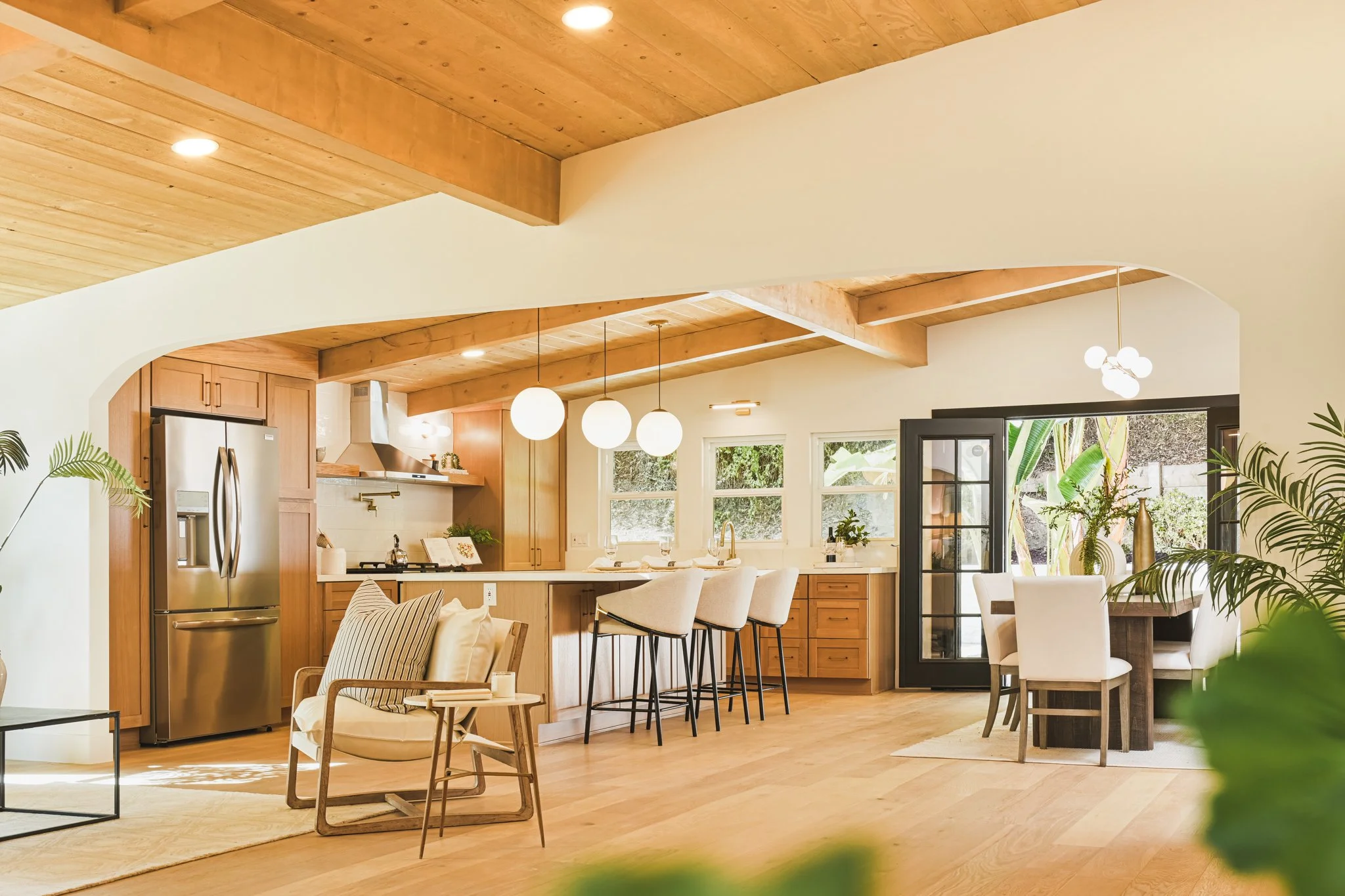 Open-concept living space with a kitchen, dining area, and seating, featuring wood accents on the ceiling and cabinetry, large windows, and modern lighting fixtures.
