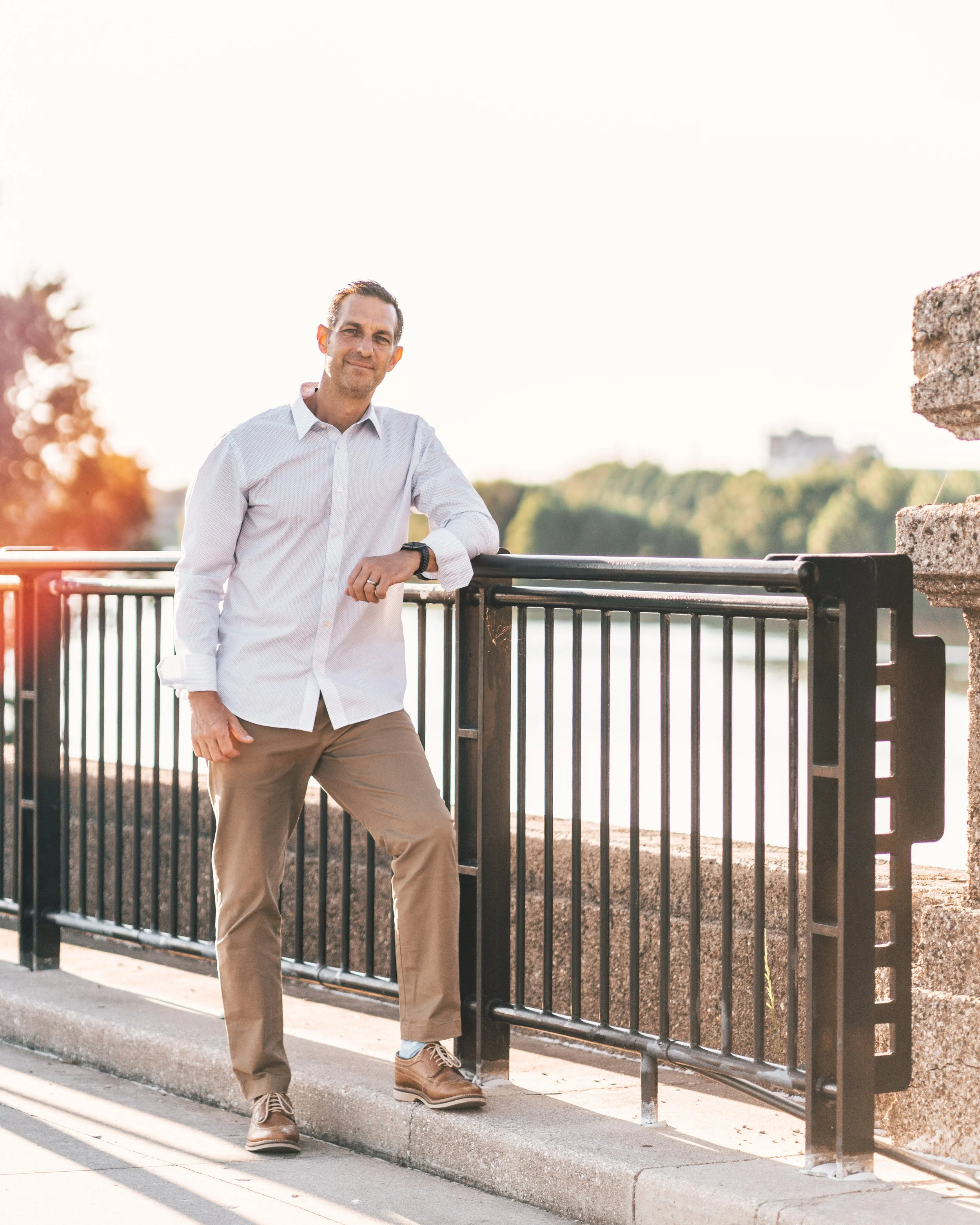 A man stands outdoors next to a black metal railing, wearing a light-colored button-up shirt, khaki pants, and brown shoes, with a relaxed expression and the sun setting in the background.