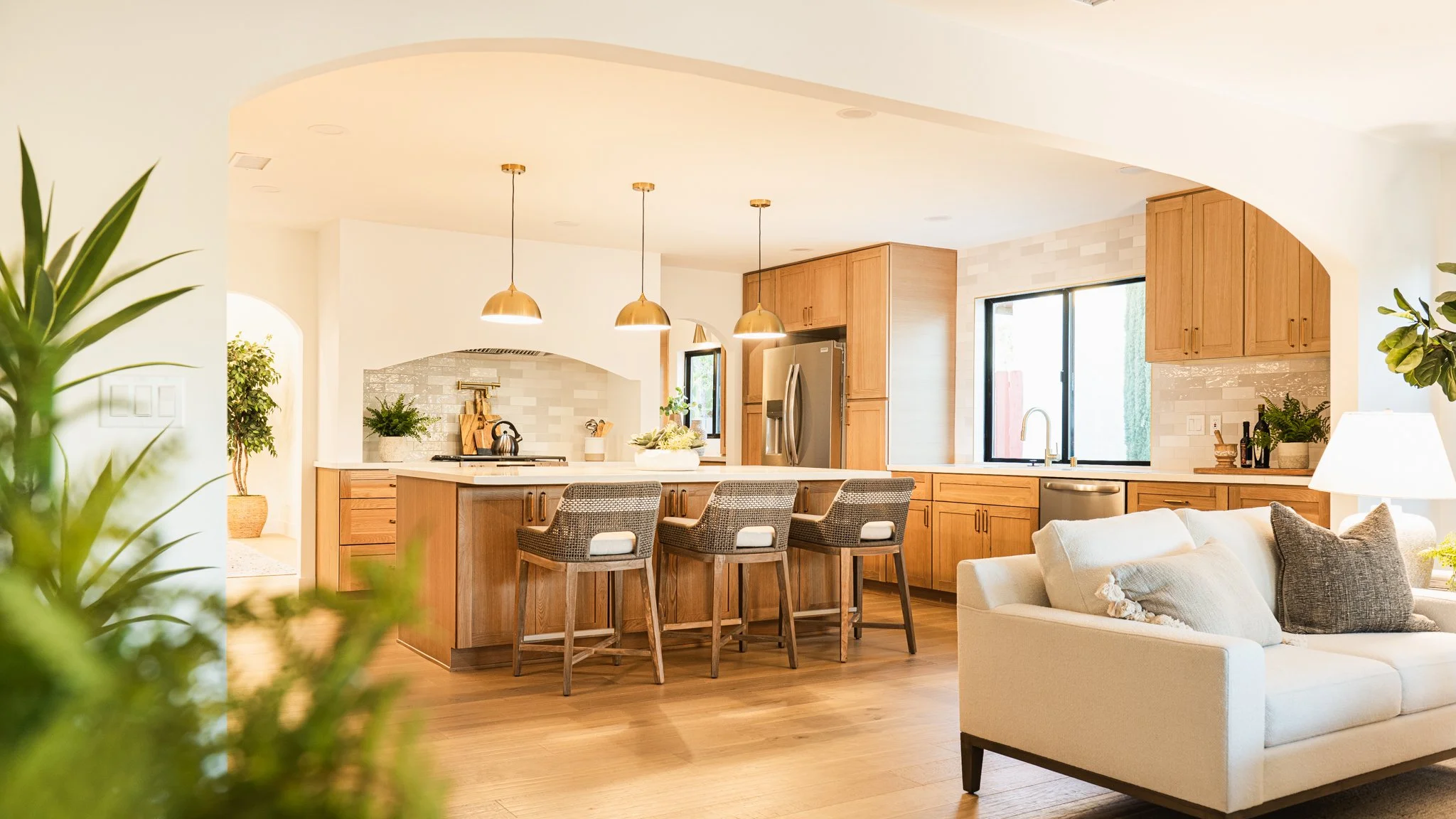 Bright and modern kitchen with wooden cabinets, a large island with bar stools, stainless steel appliances, and light fixtures hanging from the ceiling, adjacent to a cozy living area with a white sofa.