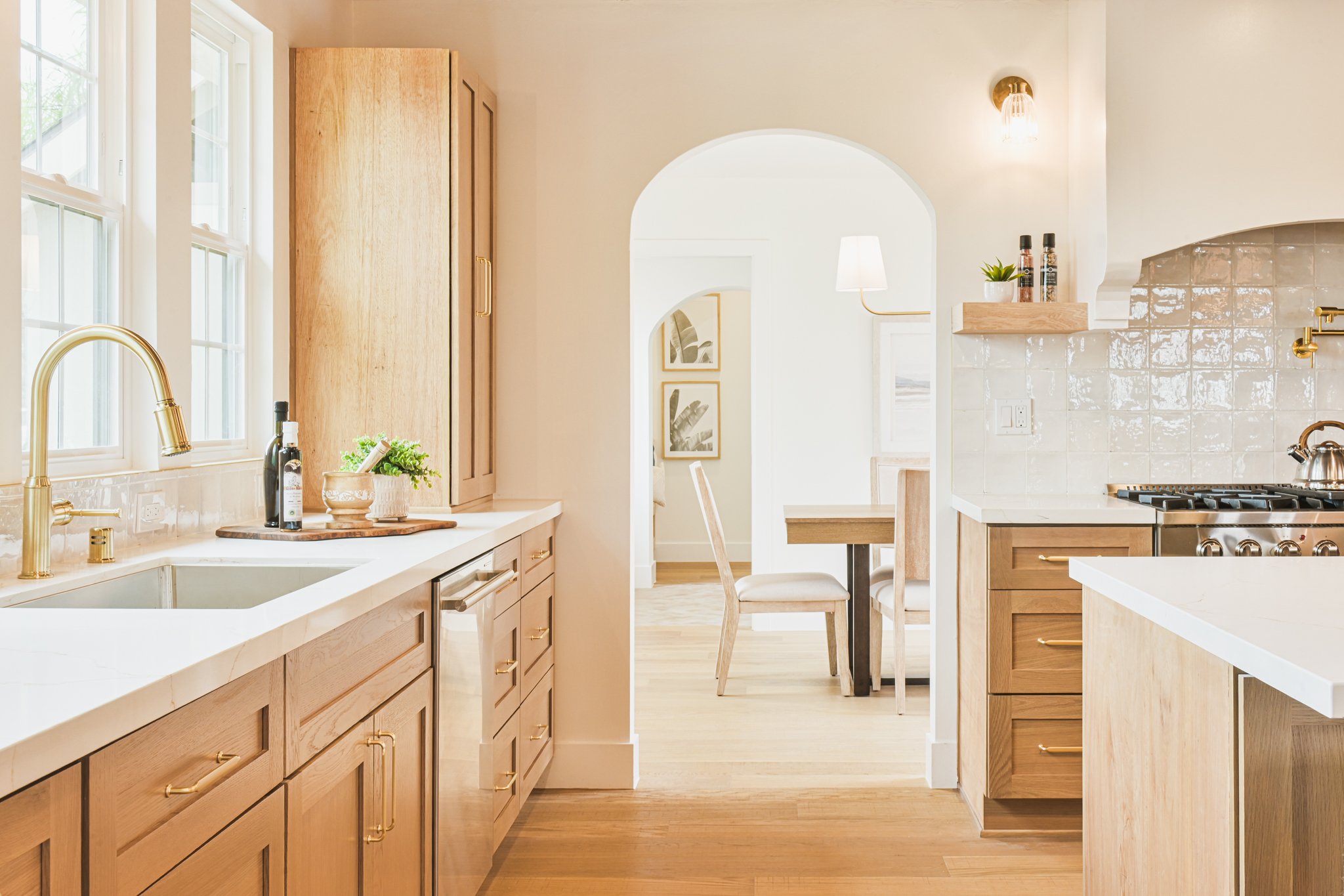 Modern kitchen with light wood cabinets, white countertops, and gold hardware, featuring a gold faucet, potted plant, and decorative jars, with a dinning area visible through an archway.