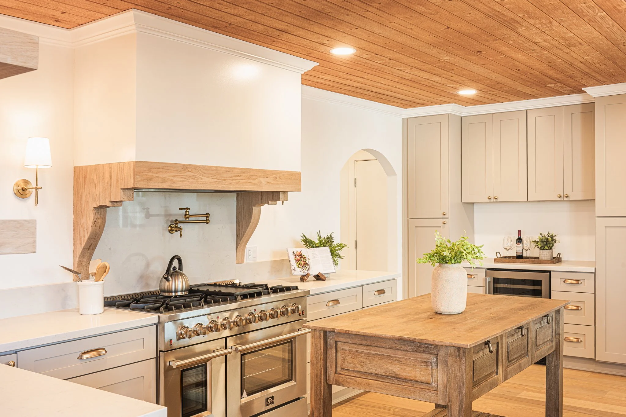 A modern kitchen with white cabinets, a wooden island, a stainless steel oven, and a wooden ceiling.