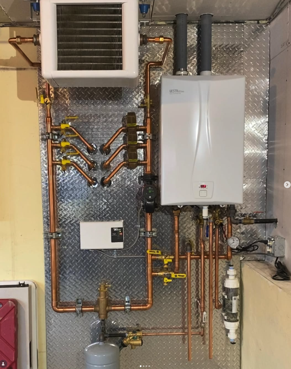 Installation of a boiler system with copper piping, valves, a water heater, and a heat exchanger on a diamond plate wall.
