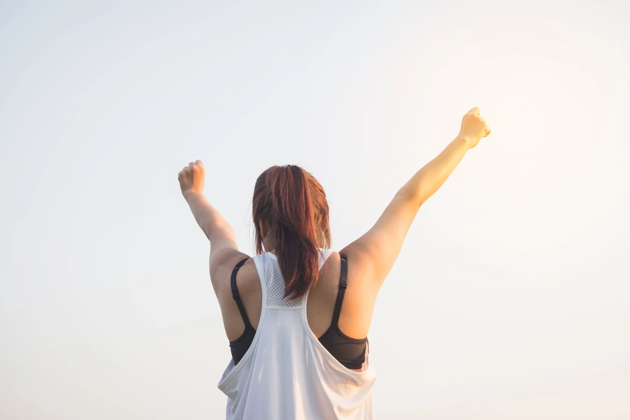 A woman with red hair wearing a white sports tank top and black sports bra, standing outdoors with her back to the camera, raising both arms in a victorious pose against a light sky.
