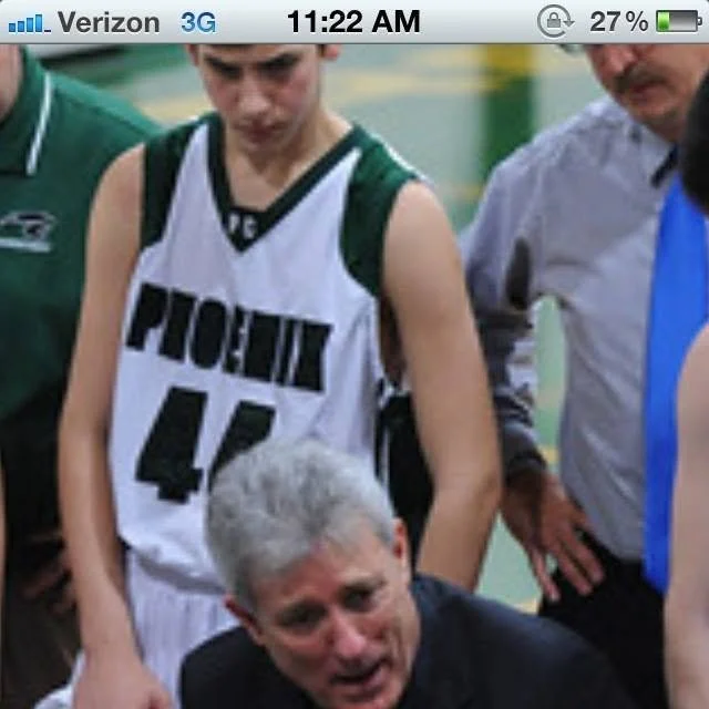 A young male basketball player in a green and white uniform with the number 45 looks down at a man with gray hair, who is speaking to or addressing the team, possibly during a game or timeout. Two other people are partially visible nearby.