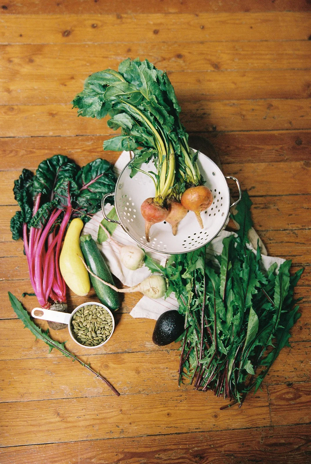 Various fresh vegetables on a wooden floor, including two turnips with greens, Swiss chard, zucchini, avocado, onion, a yellow squash, a cucumber, a scoop of pumpkin seeds, and leafy greens.