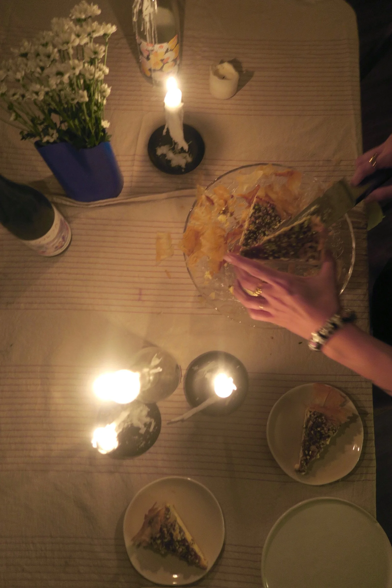 A table decorated with a lit candle, a small vase of white flowers, a glass of soda, slices of pizza, and a few empty small dishes. The table has a striped cloth. The person is holding a slice of pizza on a plate.