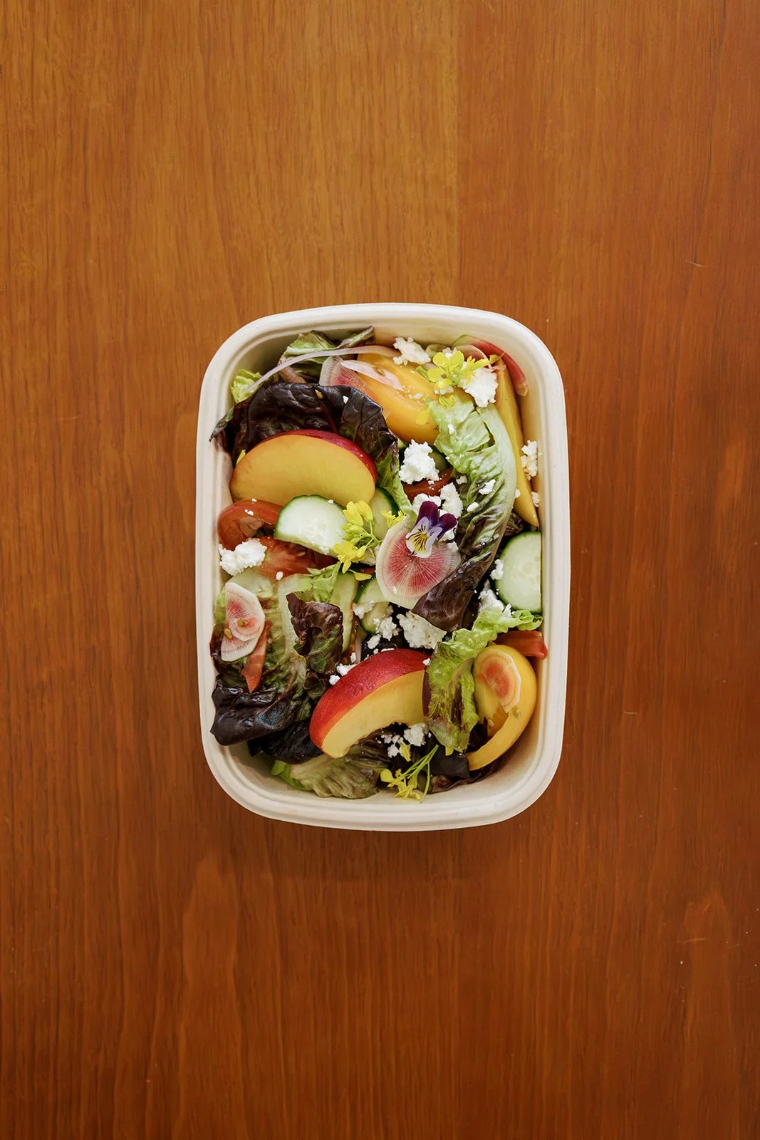 Fresh mixed salad with lettuce, cucumber, tomato, apple slices, edible flowers, and crumbled cheese in a white container on a wooden table.