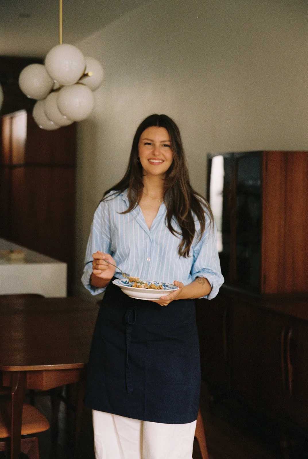 A smiling woman with long dark hair wearing a light blue striped blouse and a dark apron, holding a white plate in a cozy, well-lit room with a modern light fixture hanging from the ceiling.