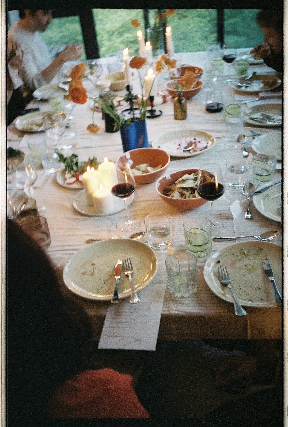 A long dining table with empty plates, glasses of water, wine, and candles. There are flowers in vases and a candlelit centerpiece. Several people are seated, some eating and some looking at their phones. The background shows large windows with a view of greenery outside.