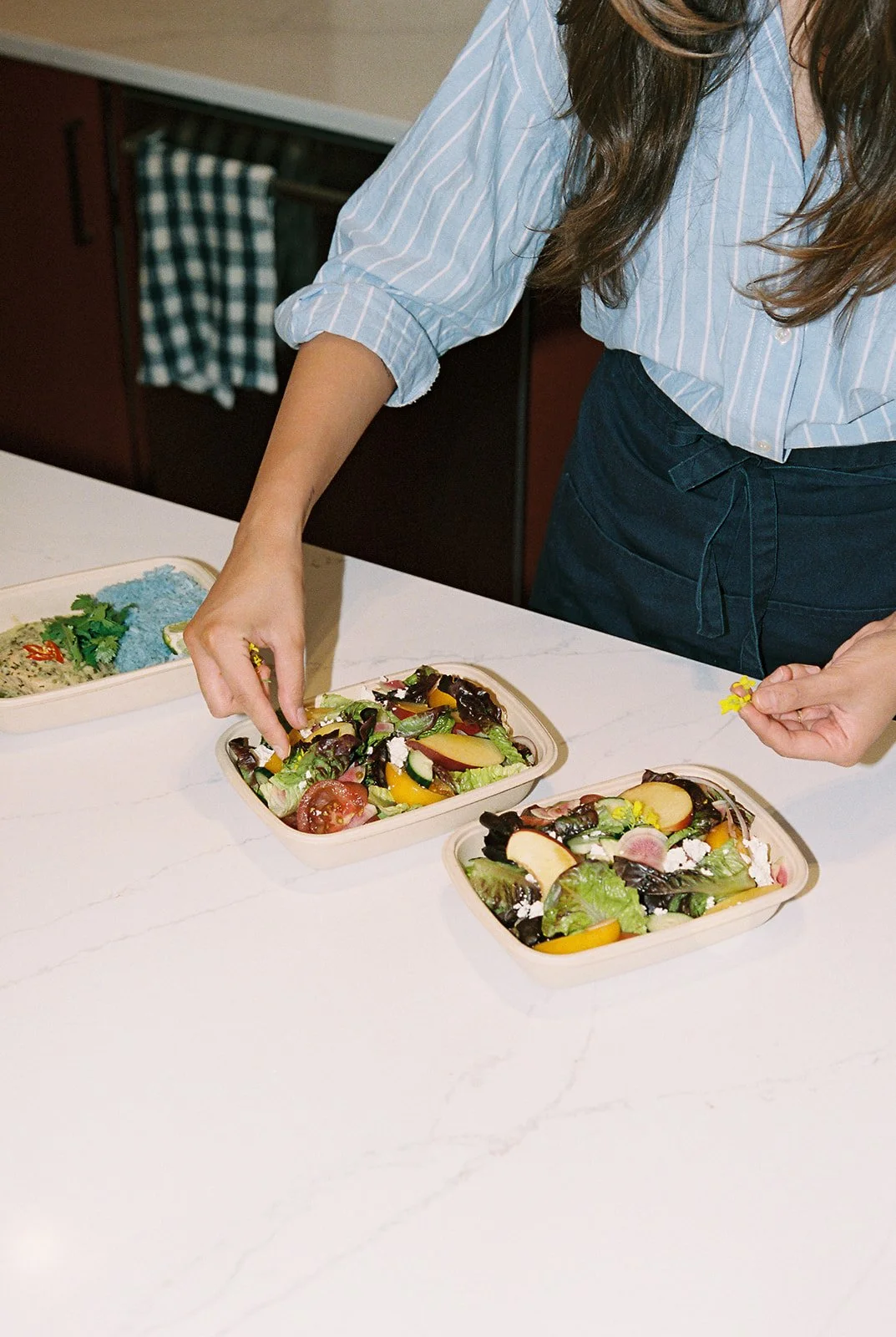 Person preparing salads in takeaway containers on a white kitchen counter.