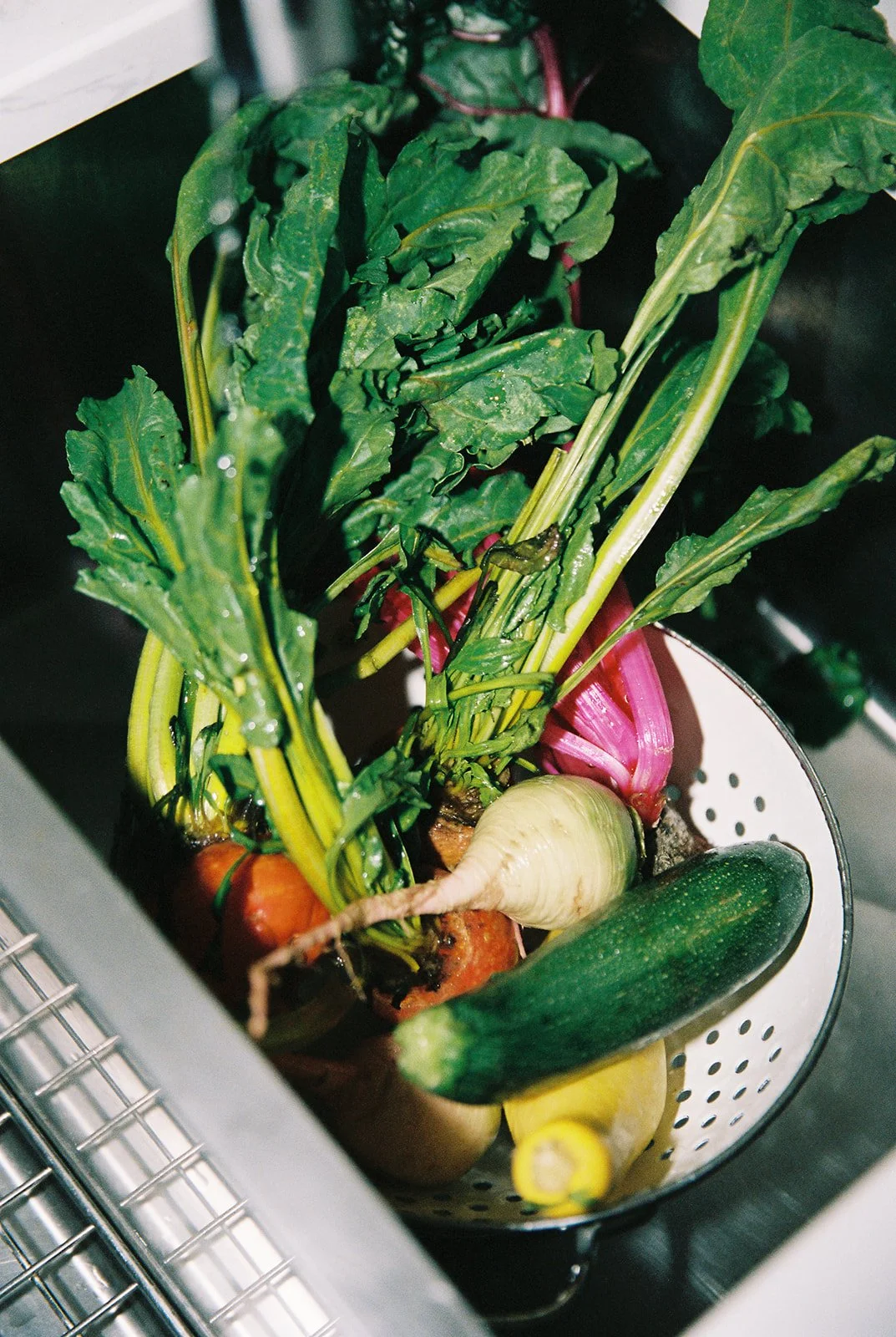 Fresh vegetables including a zucchini, turnip, beets with greens, and a cucumber inside a colander or strainer.