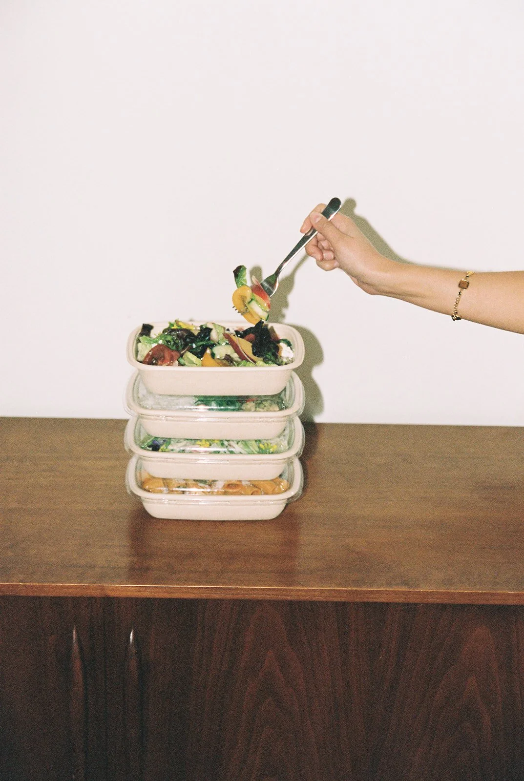A hand with a gold bracelet is scooping salad from a stack of four glass meal prep containers on a wooden table against a plain white wall.