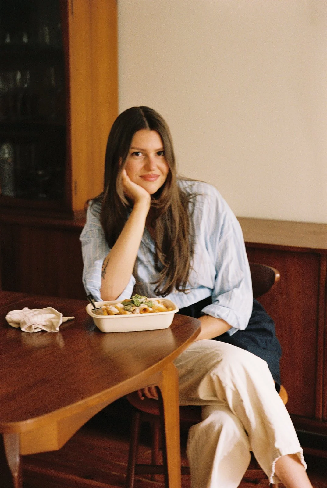 A woman with long brown hair, wearing a light blue striped shirt and beige pants, sitting at a wooden dining table with a bowl of pasta in front of her and her hand resting on her chin, in a cozy dining room.