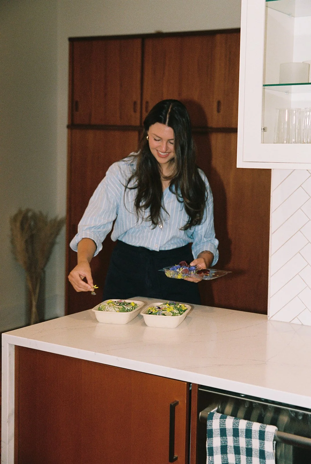 A woman with long dark hair, wearing a blue and white striped blouse, is smiling and preparing salads in a modern kitchen.