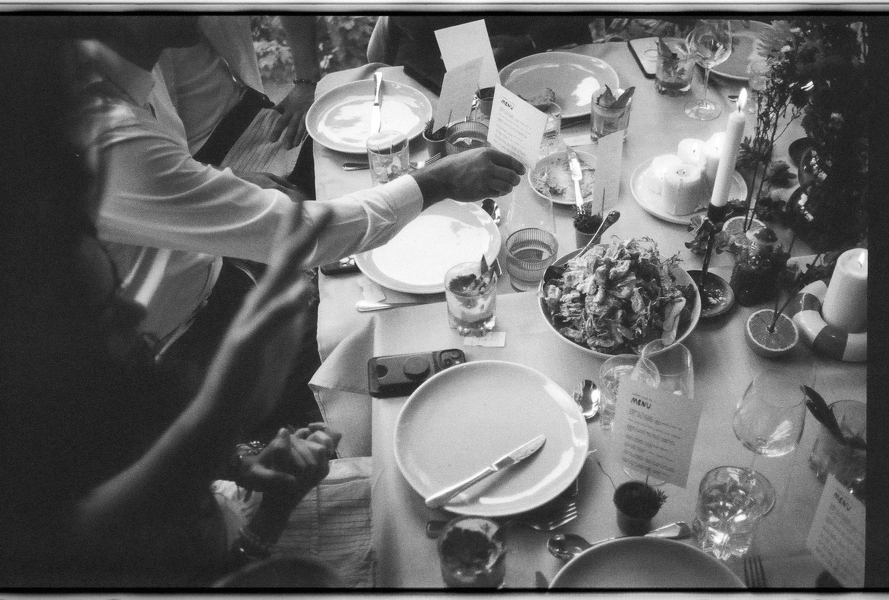 Table set for a meal with plates, glasses, cutlery, a menu, candles, a large bowl of food, and decorated with flowers and citrus slices.