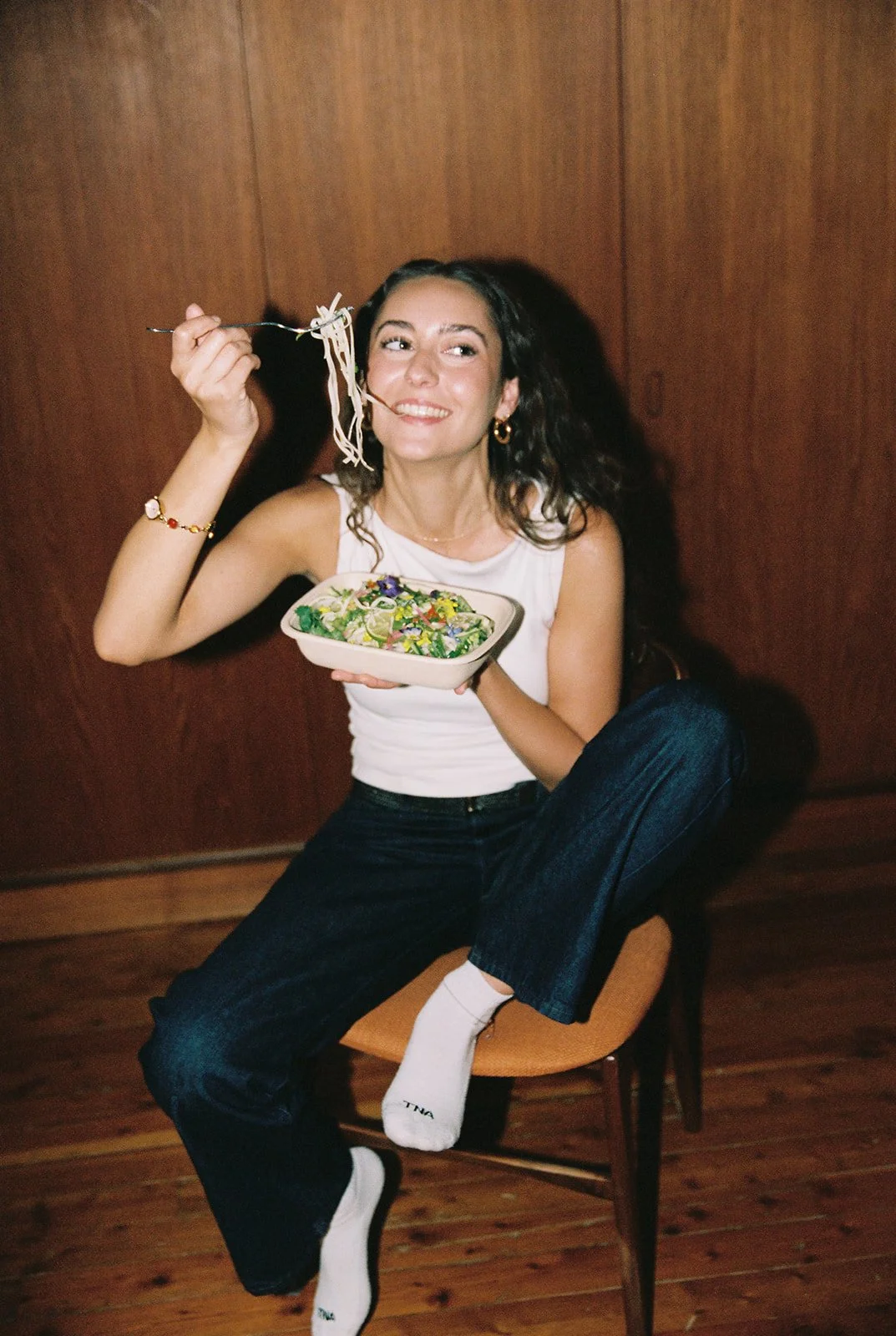 A woman sitting on a chair, holding a bowl of salad in one hand and a fork with noodles in the other, smiling and looking to her right, against a wooden wall background.