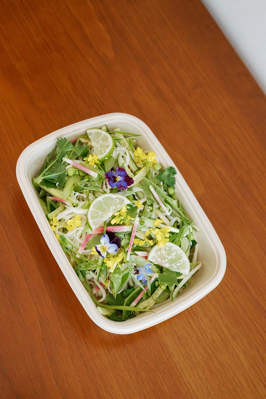 A rectangular container filled with a fresh salad of sliced cucumbers, lime wedges, edible flowers, and herbs on a wooden surface.