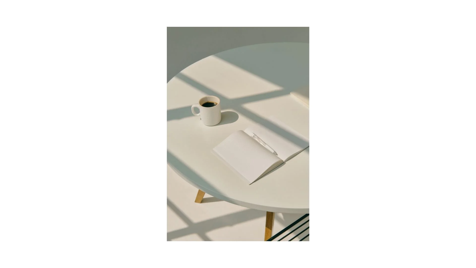 A white round table with wooden legs holding an open notebook and a pen, a white coffee cup filled with black coffee, under a window with sunlight casting shadows.
