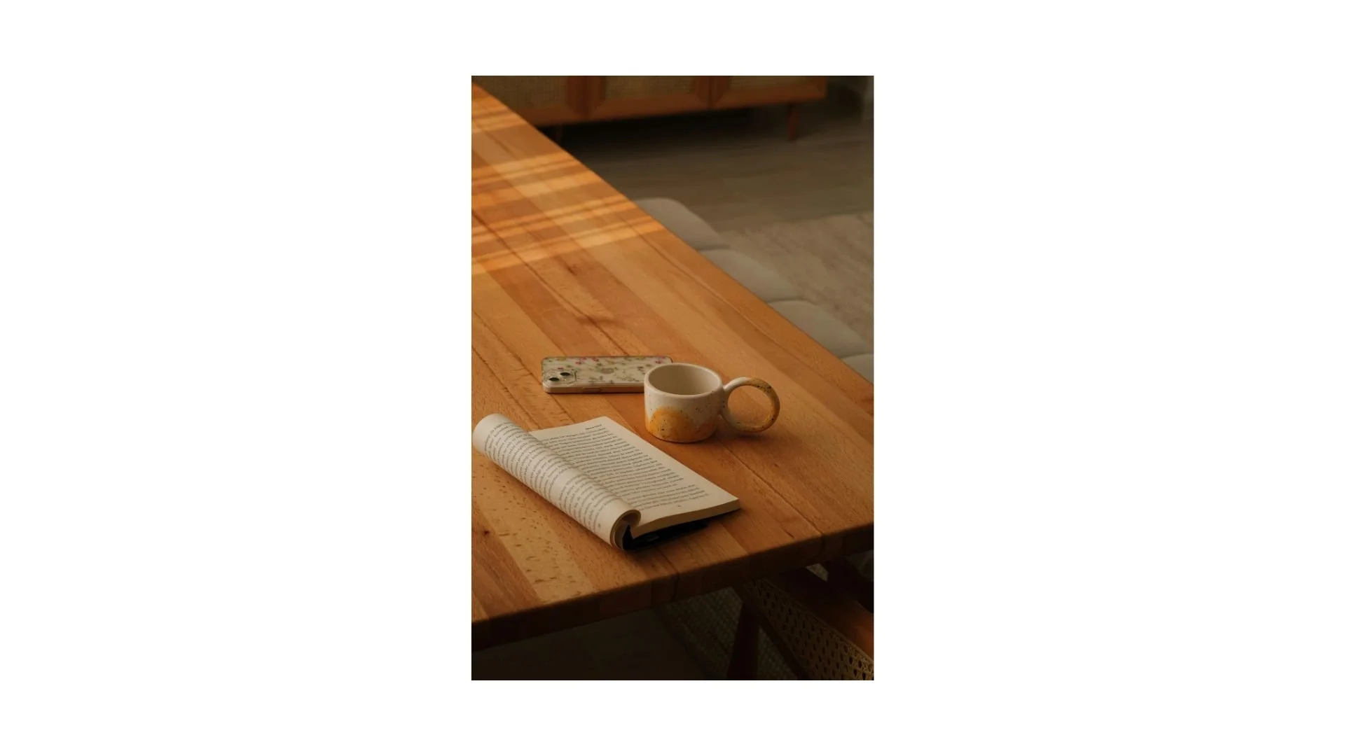 A wooden table with a book, a ceramic mug, a smartphone, and a rolled-up newspaper.