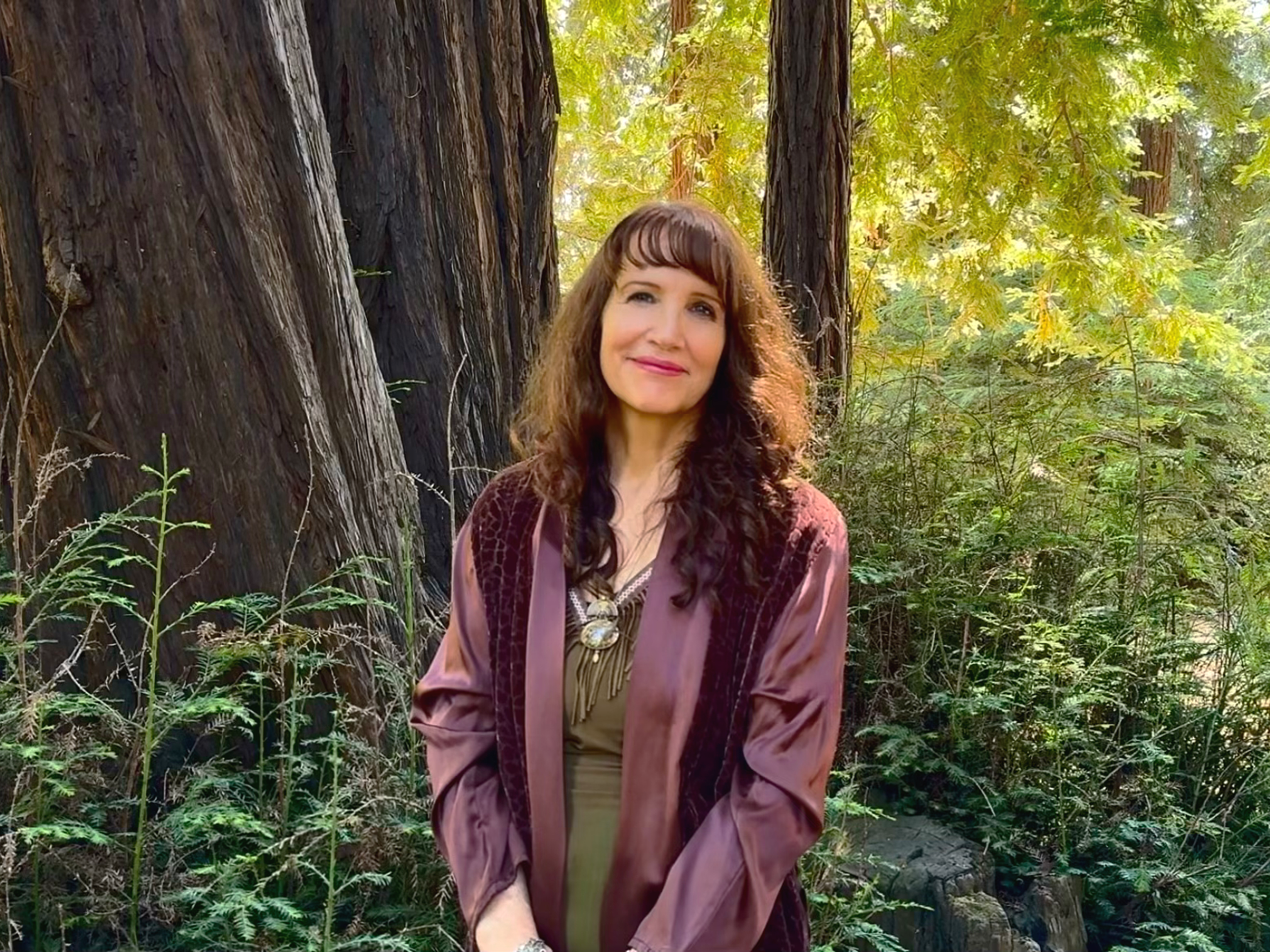 A woman with long brown curly hair, wearing a mauve jacket and beige top, standing outdoors in a forest with large trees and green foliage.