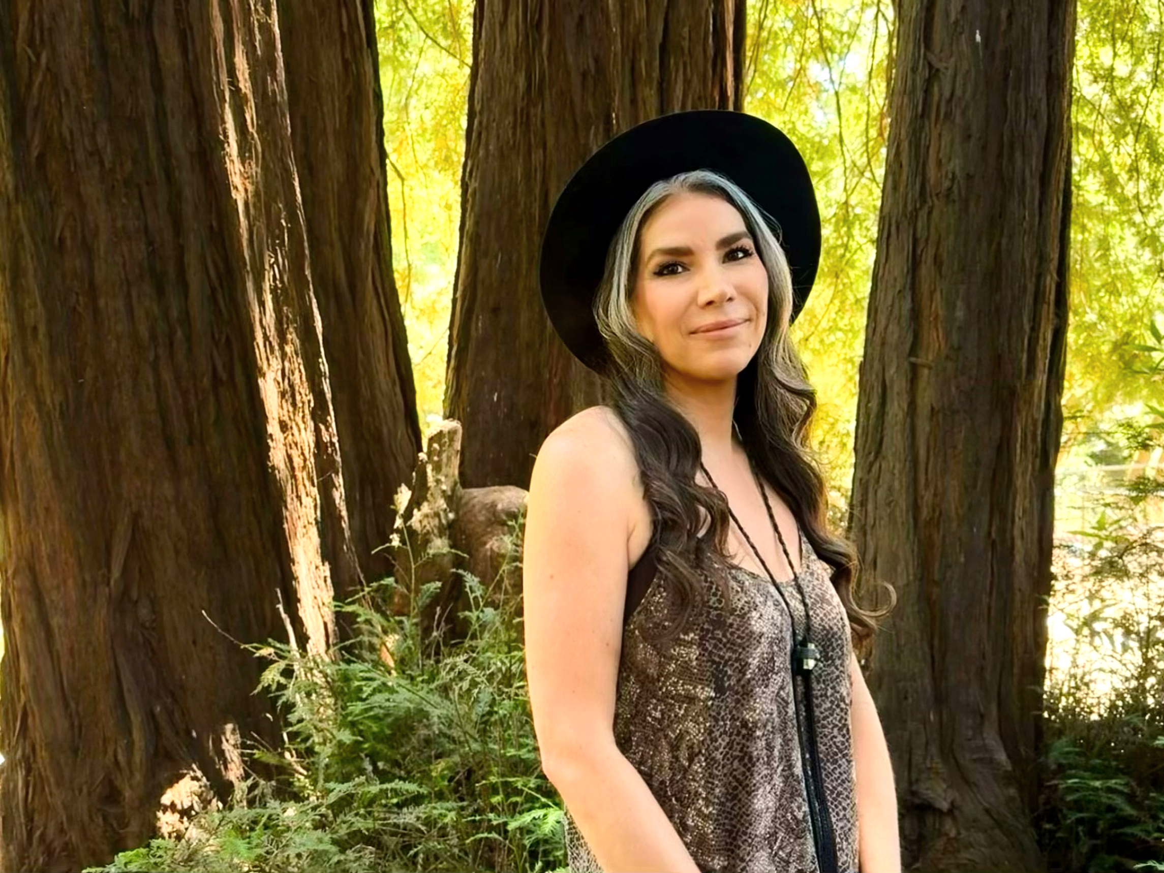 A woman with long wavy hair, wearing a black hat and a sleeveless patterned dress, standing outdoors in front of tall trees with green foliage.