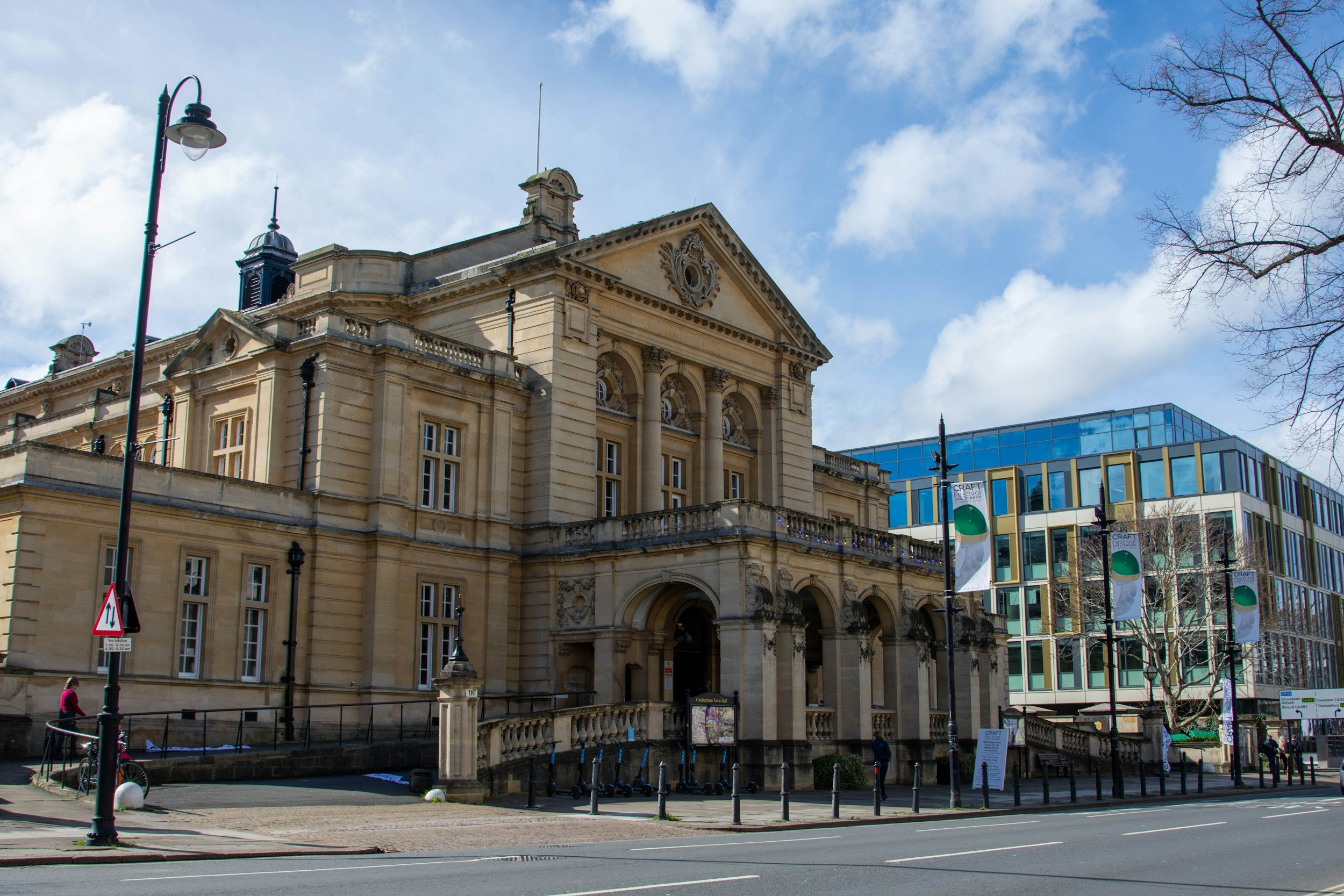 A historic stone building with ornate architectural details and a triangular pediment, adjacent to a modern glass structure, under a partly cloudy sky, with trees and street lamps in the foreground.