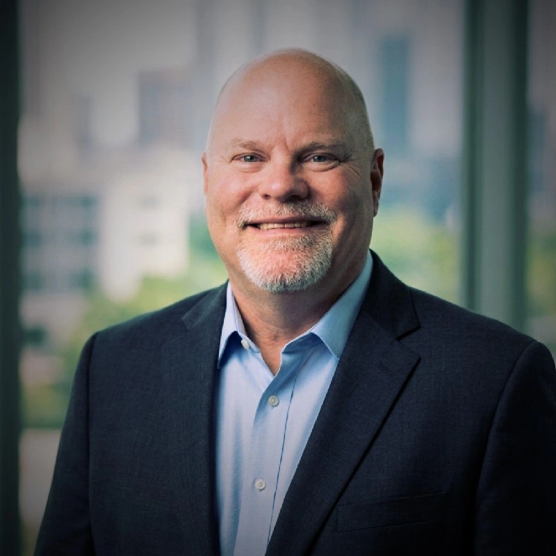 Portrait of a middle-aged man with a bald head and a beard, wearing a dark suit jacket and a light blue dress shirt, smiling in an office setting with glass windows and blurred cityscape background.