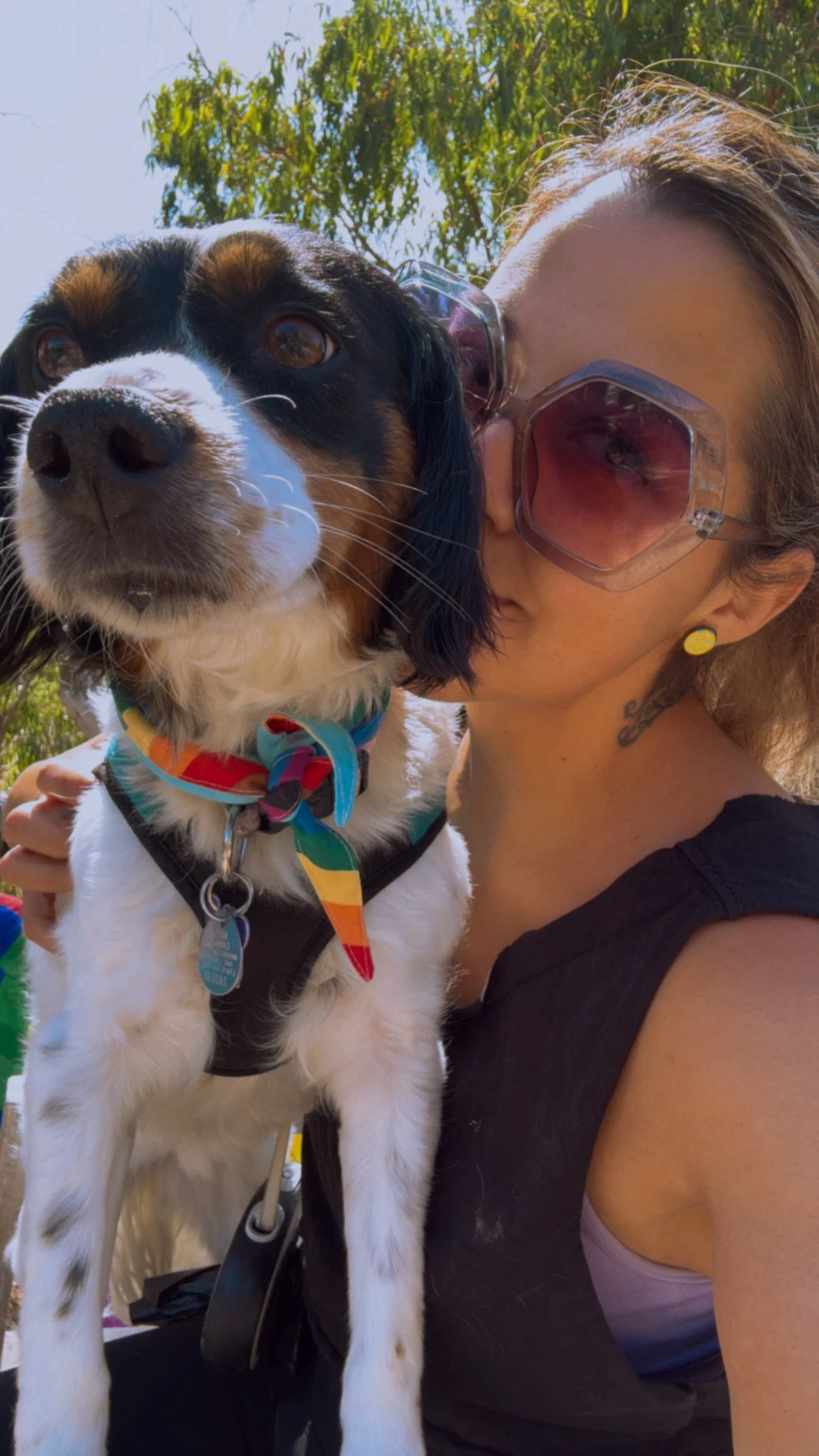 Woman wearing pink sunglasses and a black tank top holding a black and white dog outdoors on a sunny day.