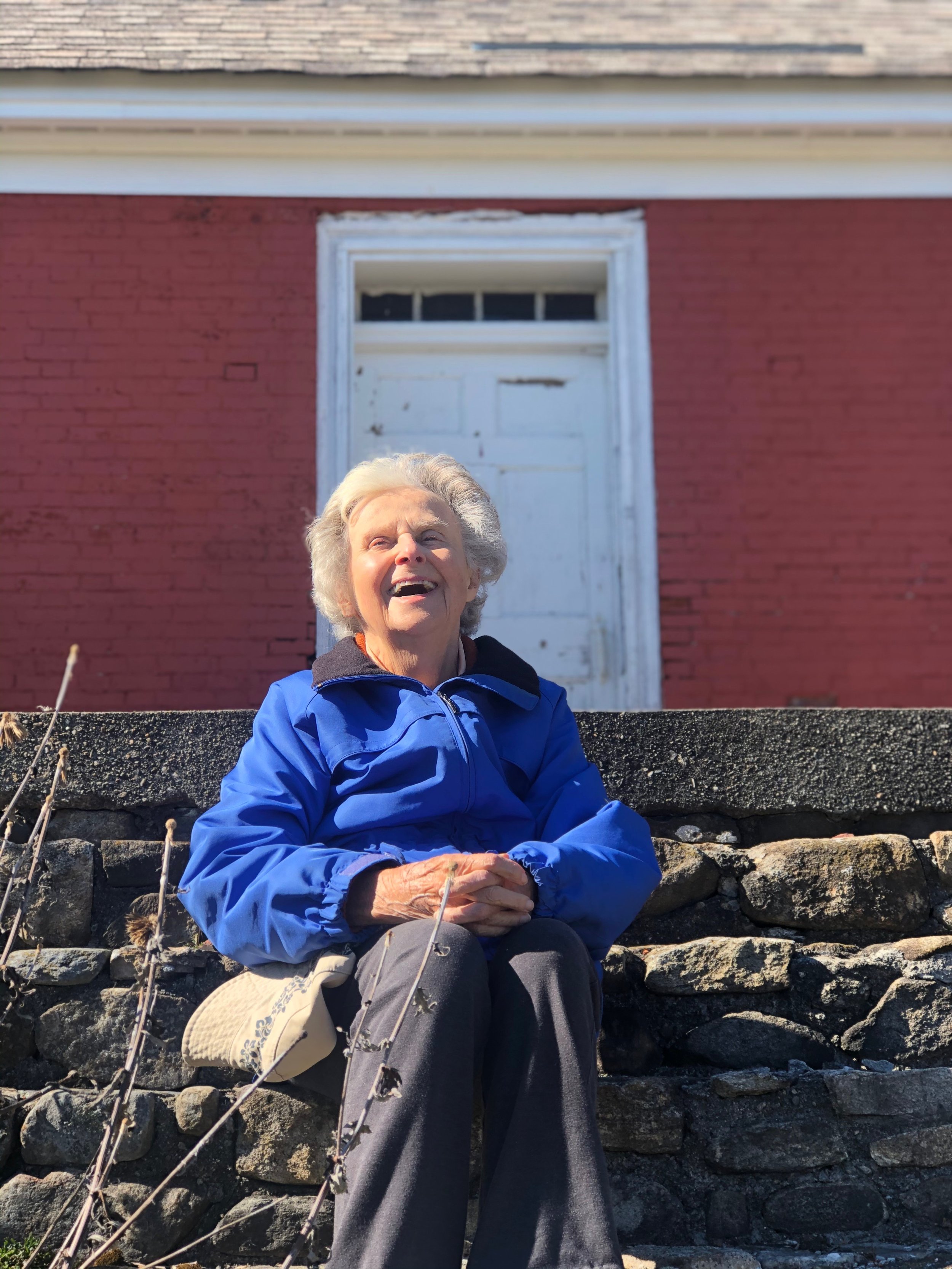 An elderly woman with gray hair sitting outdoors on stone steps smiling, with a red brick building and a closed white door in the background.