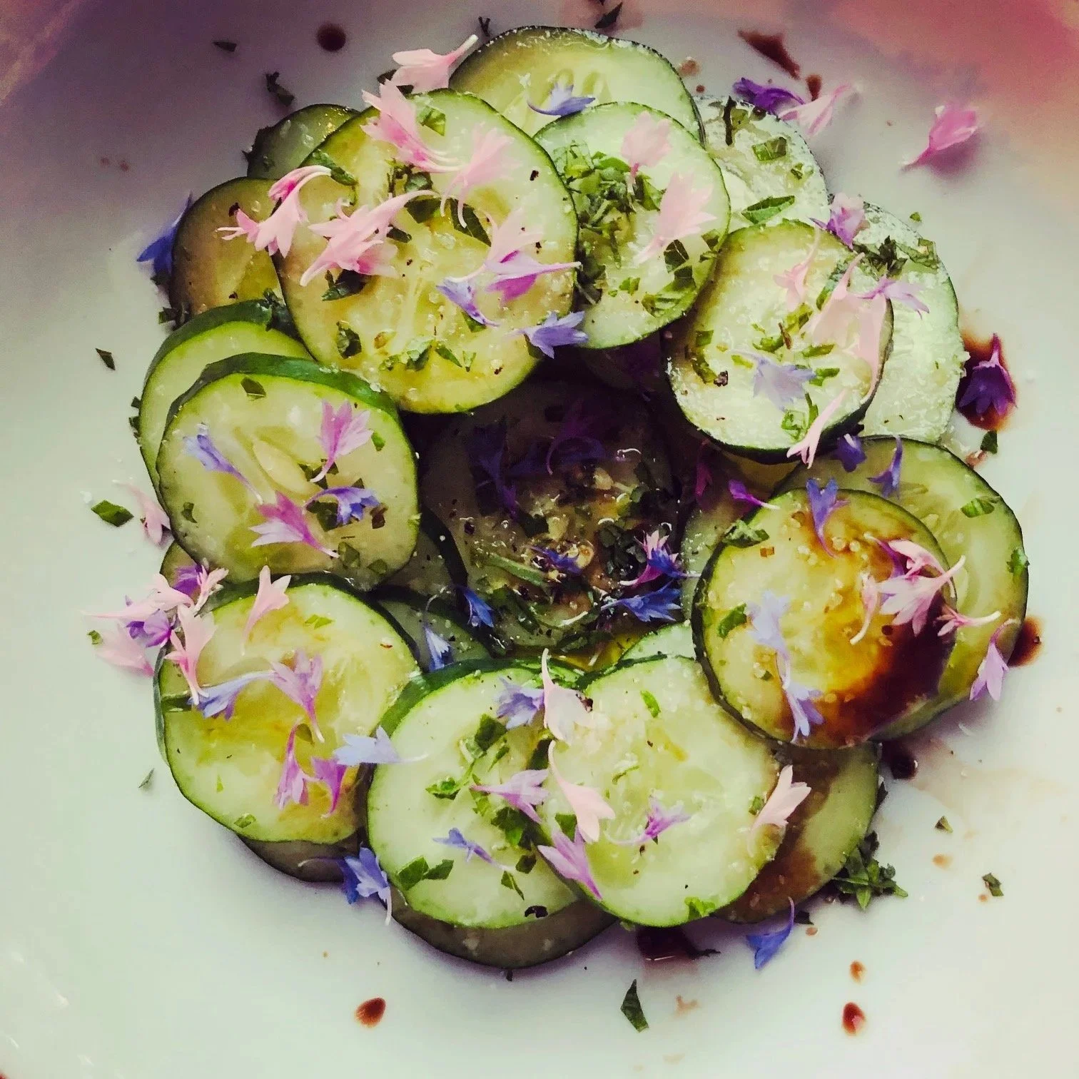 Sliced cucumbers garnished with edible pink and purple flowers and chopped herbs, with a drizzle of sauce on a white plate.
