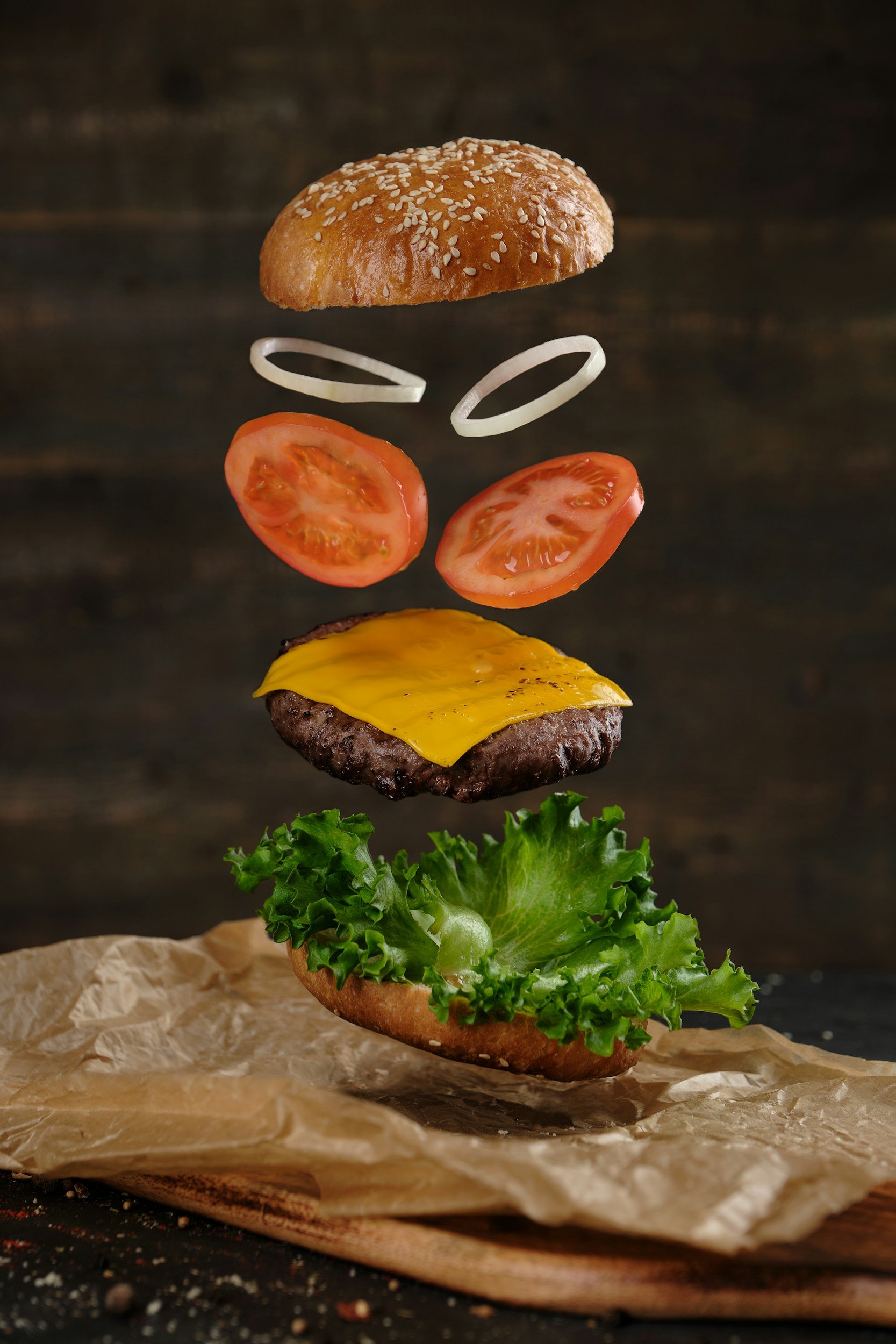 A hamburger with a sesame seed bun, lettuce, a cheeseburger patty, tomato slices, onion rings, and a hamburger bun, with ingredients appearing to float mid-air above a crinkled paper on a dark surface.