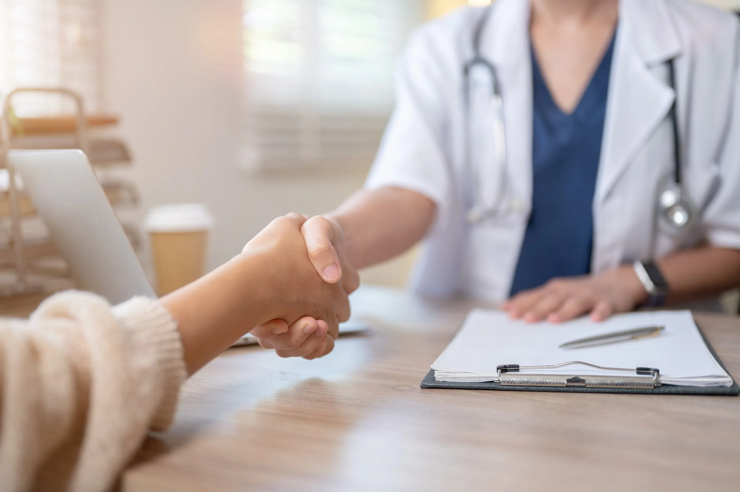A healthcare professional in a white coat and stethoscope shaking hands with a patient in a cozy, well-lit office or clinic.