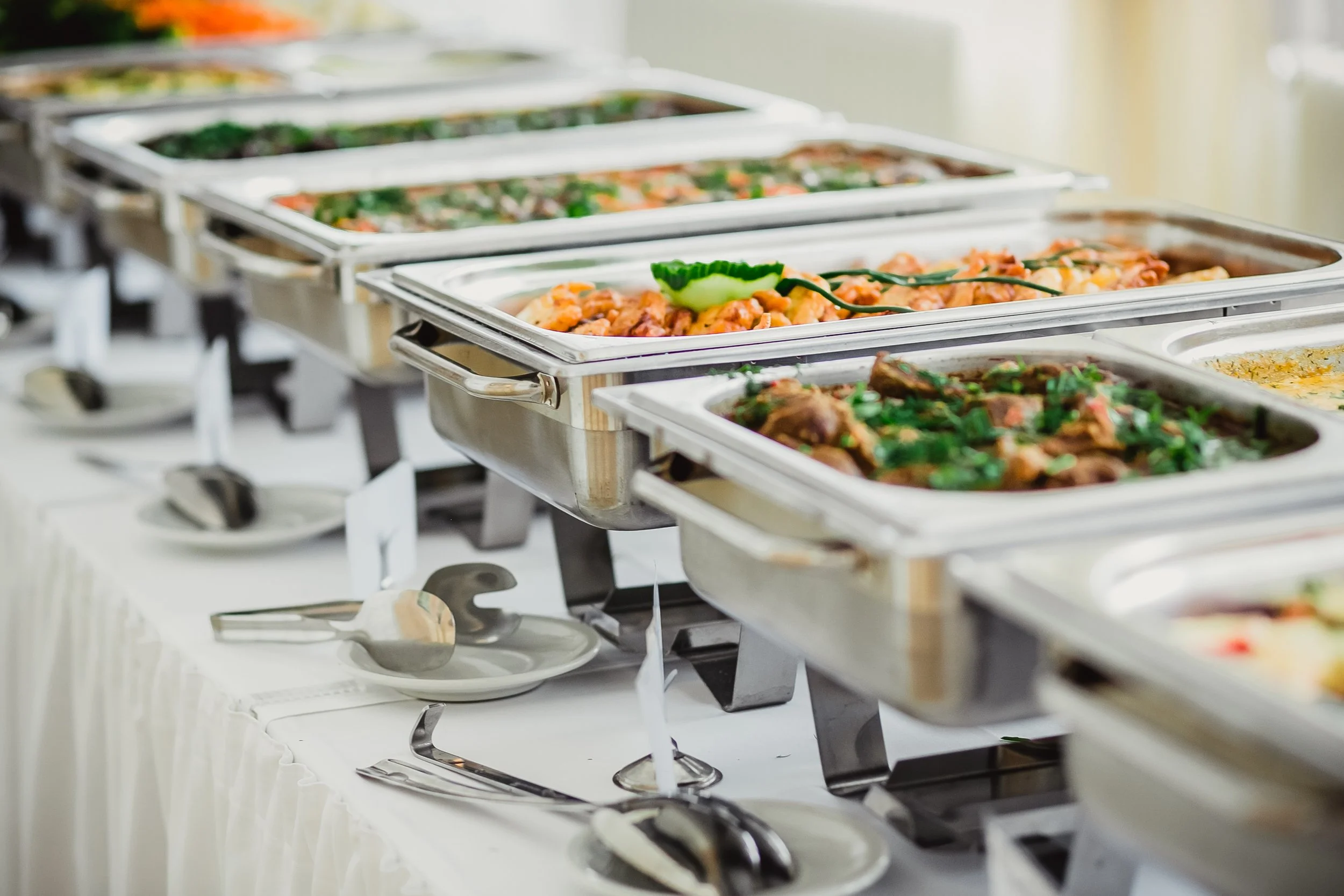 Buffet table with stainless steel chafing dishes filled with various hot food items, garnished with vegetables, on a white tablecloth at a dining event.
