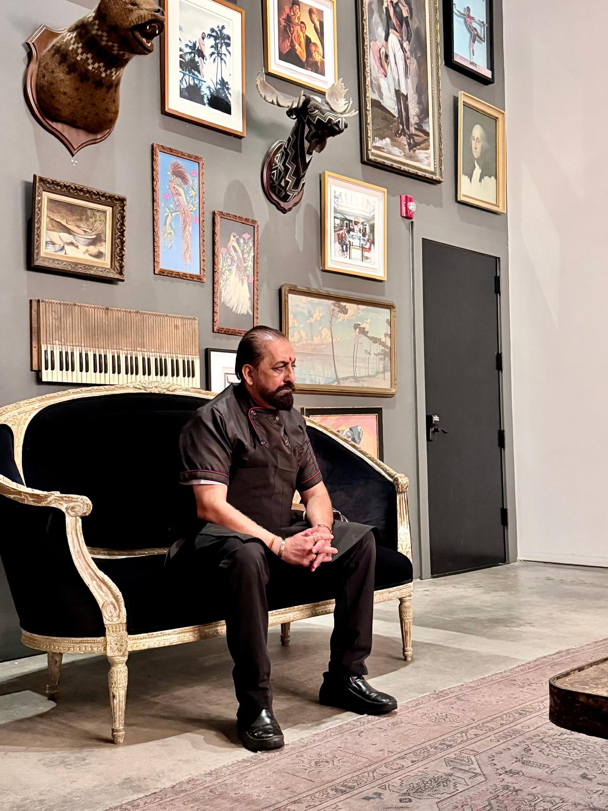 A man with a beard and long hair tied back, wearing a black uniform and sitting on a vintage black and gold sofa, in a room with gray walls. The wall behind him is decorated with a variety of framed pictures, paintings, and mounted animal heads, including a bear and a deer.