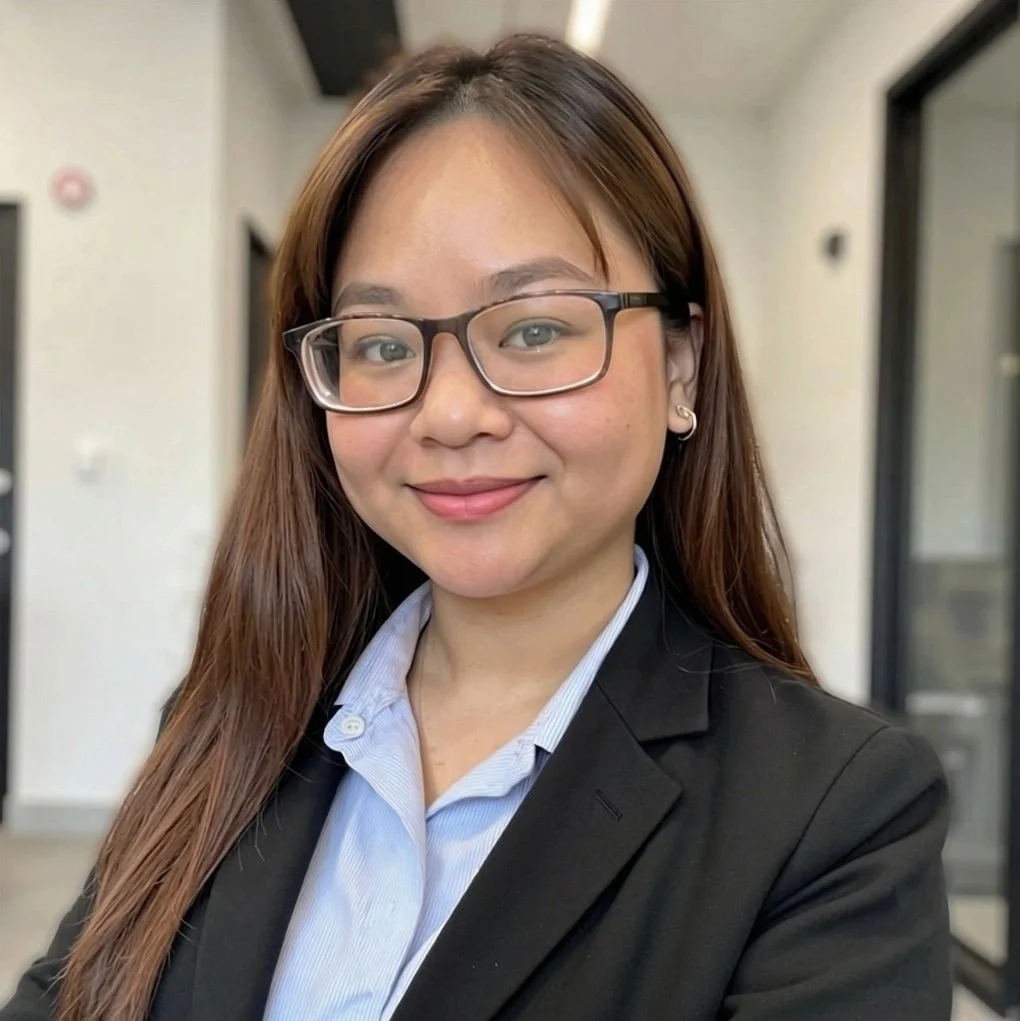 A portrait of a woman with long brown hair, wearing glasses, a black blazer, and a light blue shirt, smiling in an indoor setting.