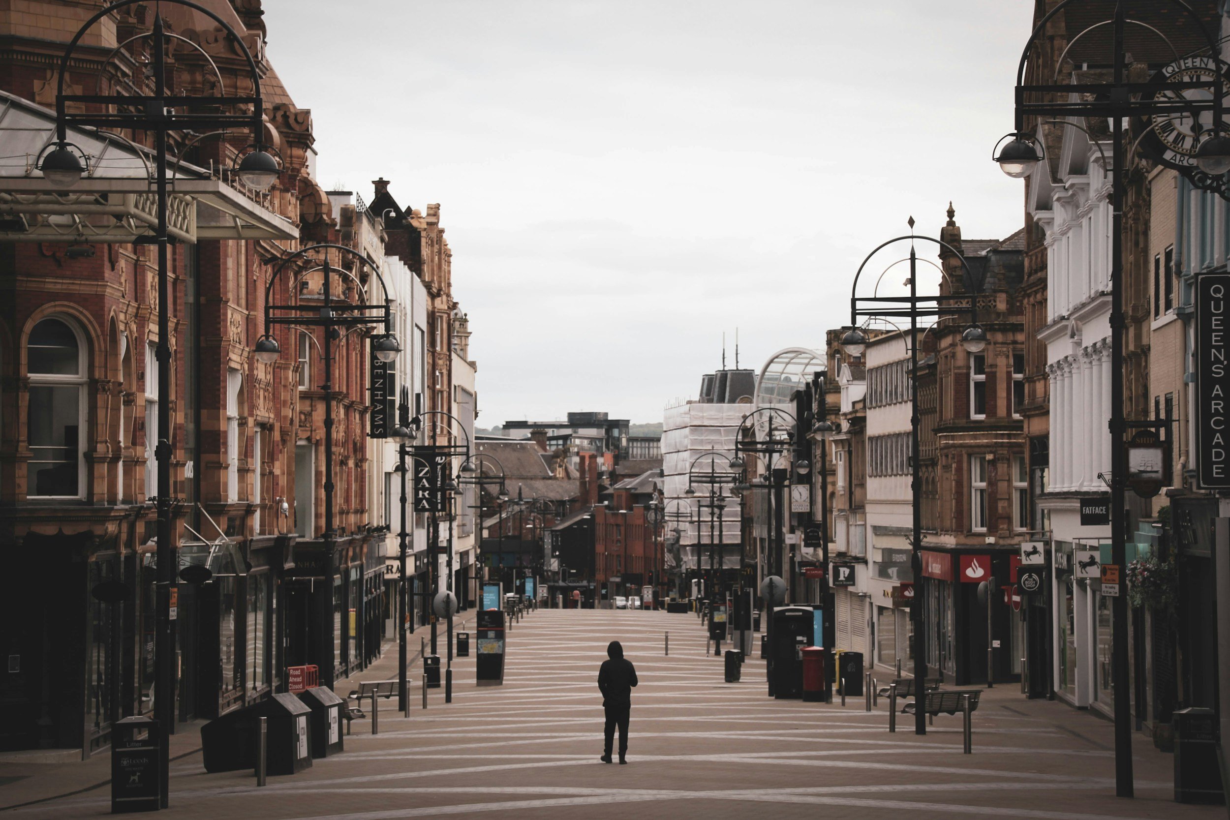 An empty city street with historic brick and white buildings, street lamps, and a person walking in the distance on a cloudy day.