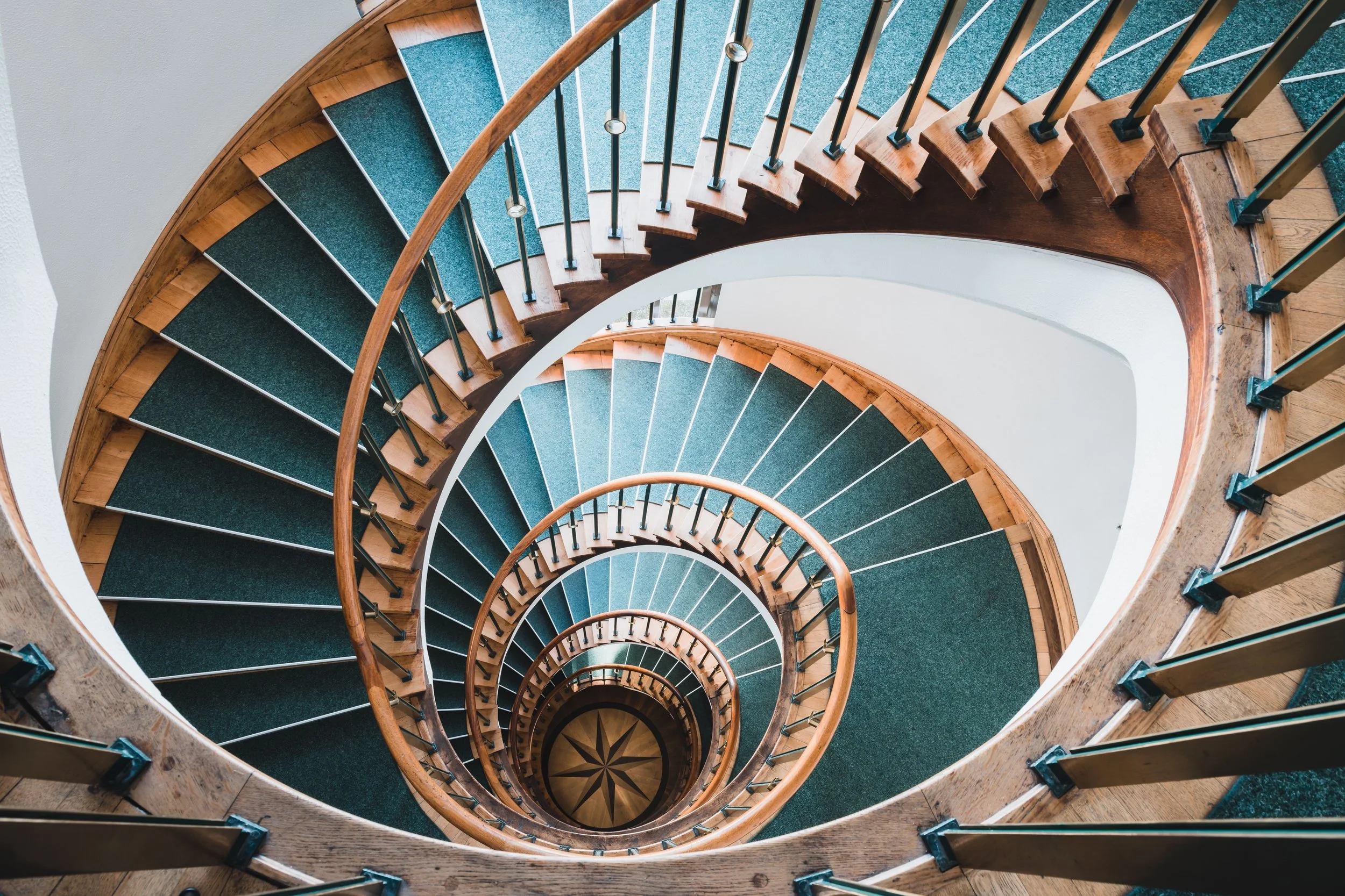 Top-down view of a spiral staircase with wooden steps, a green carpet runner, metal railings, and a wooden handrail.