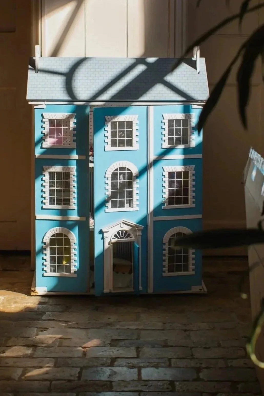 A dollhouse modeled after a blue, three-story mansion with white trim, multiple windows, and a front door with a decorative arch, inside a room with sunlight casting shadows.
