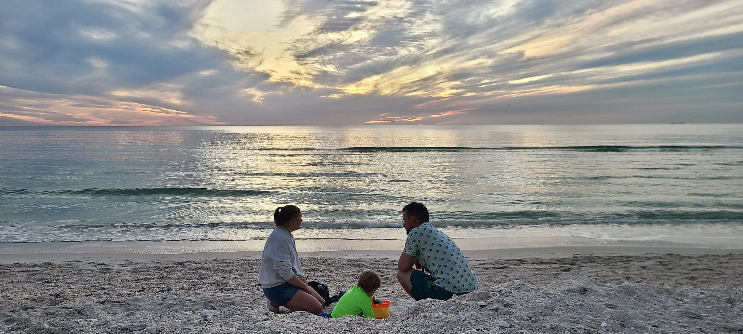 A family of three spending time together on a beach at sunset. The parents and child sit on the sandy shore, with the ocean and colorful sky in the background.