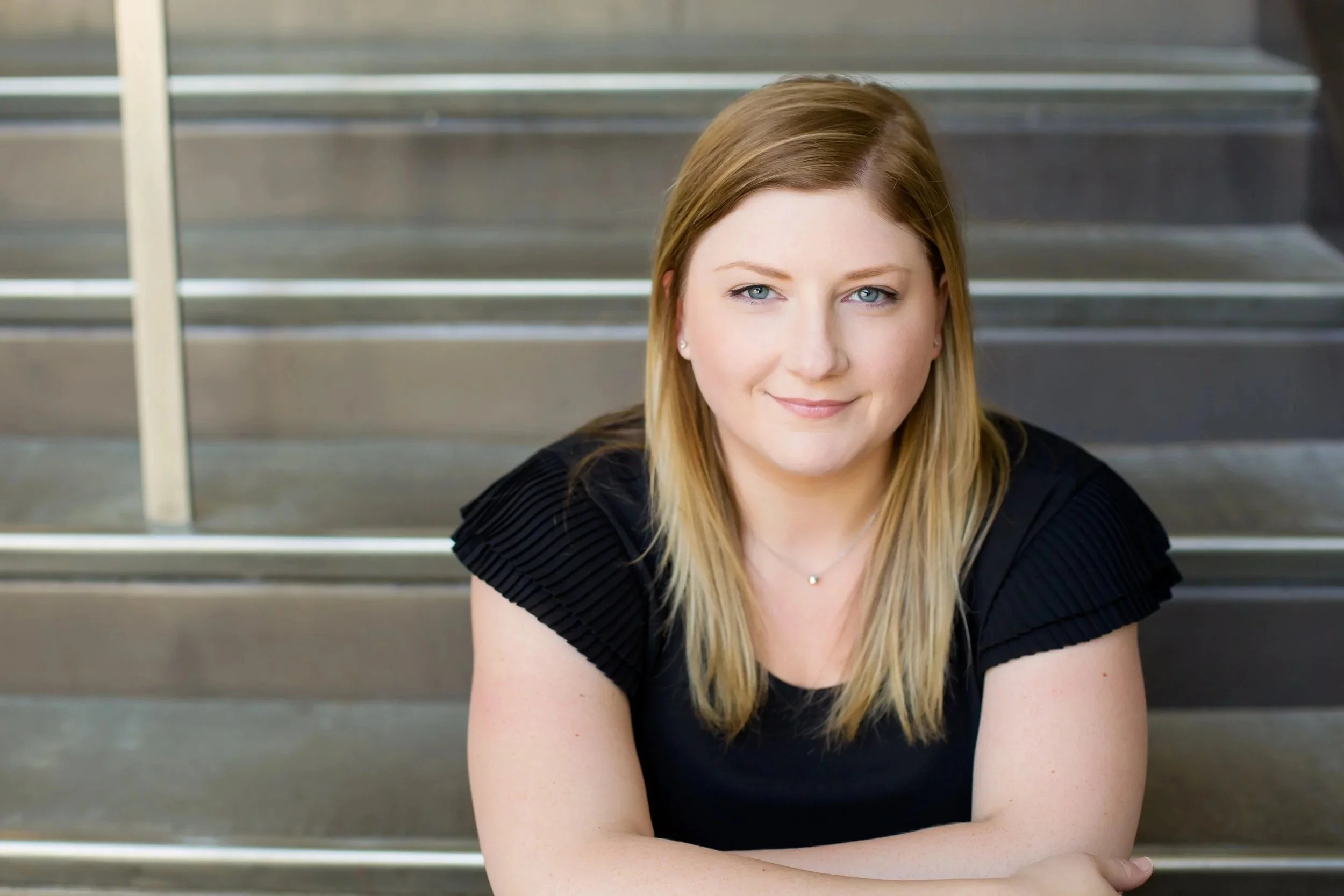 A young woman with straight blonde hair and blue eyes smiling while sitting on outdoor concrete stairs, wearing a black short-sleeved top.