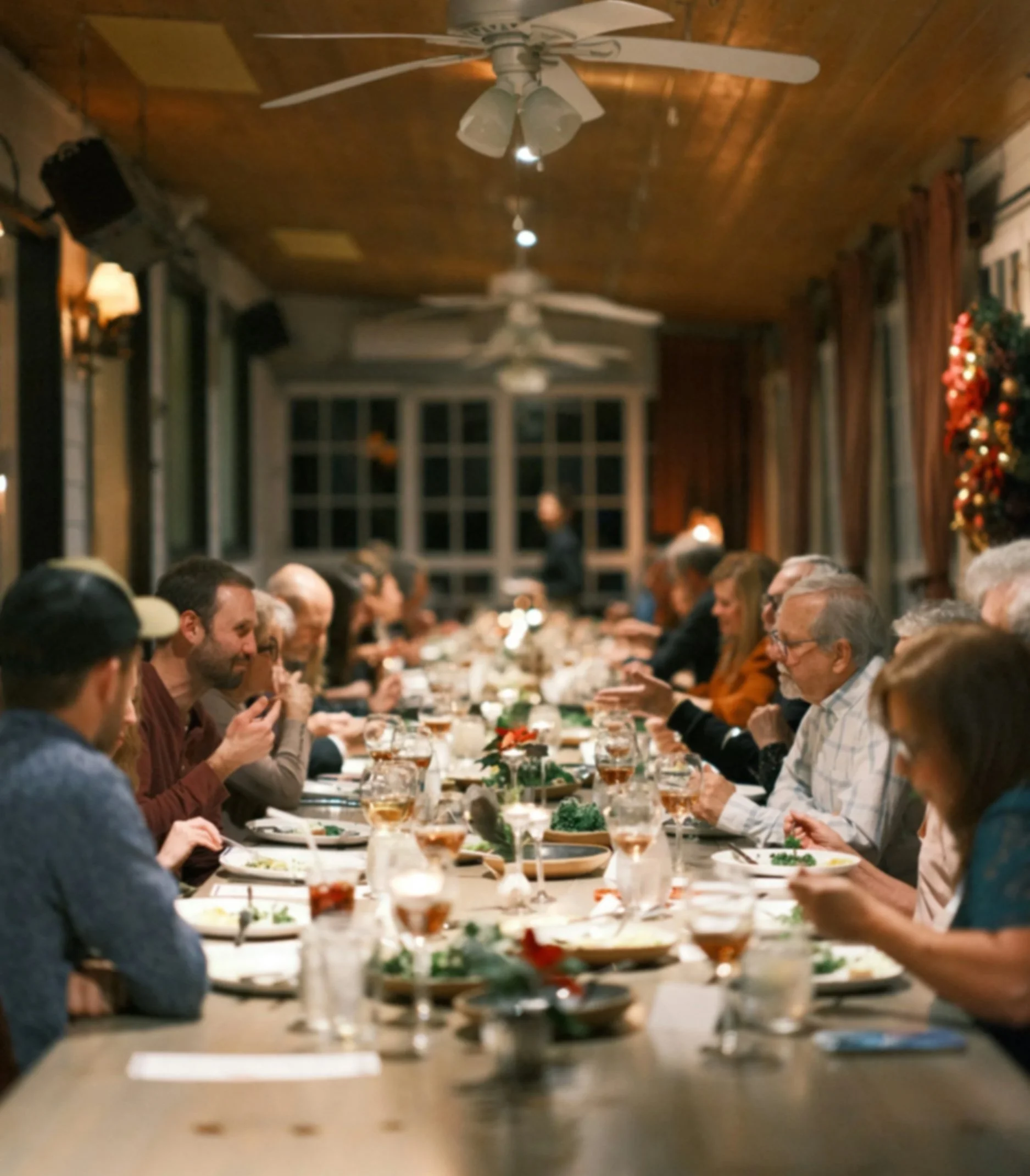 Dinner party with people seated at a long table set with glasses, plates, and salads in a warmly lit room with wooden ceiling and large windows.