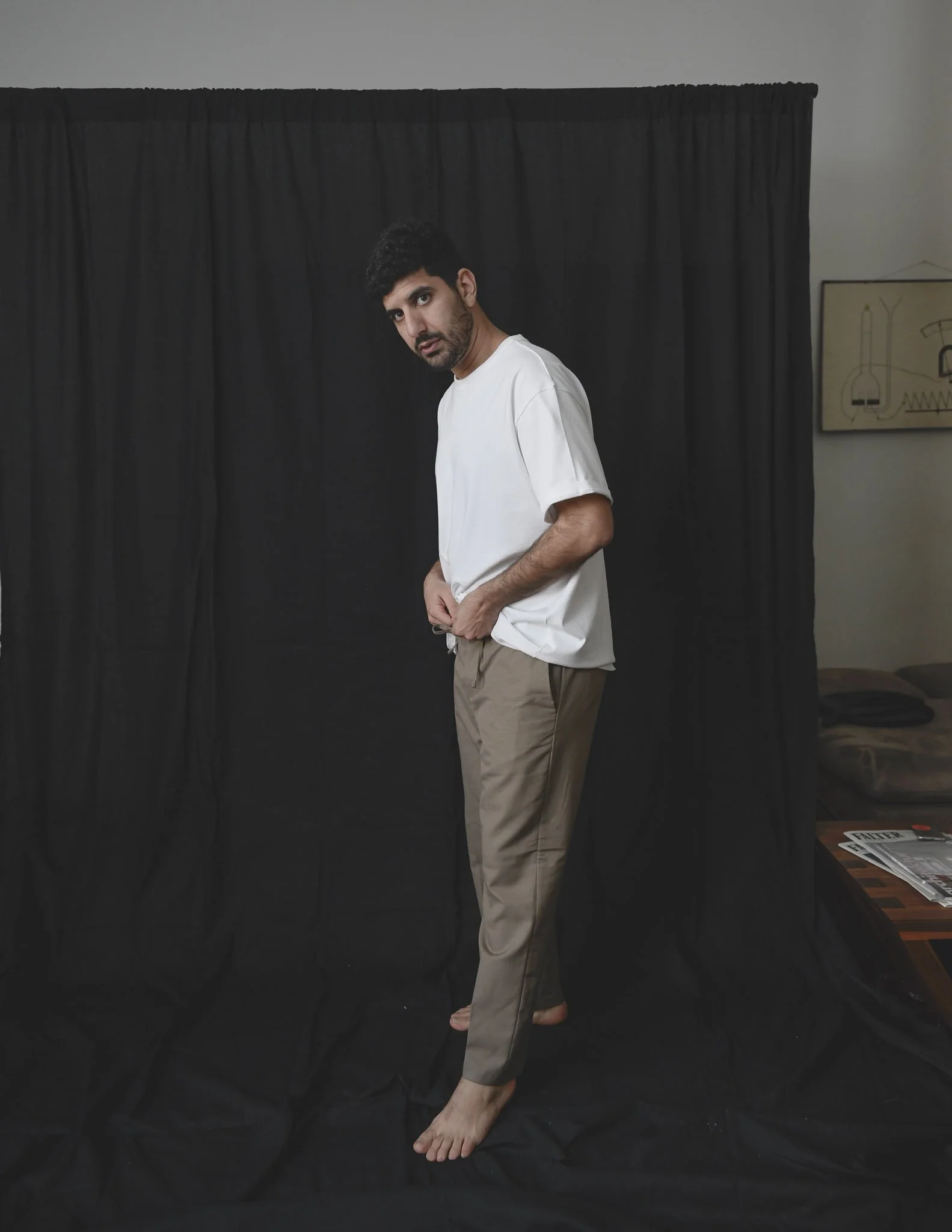 Ein Mann steht vor einem schwarzen Vorhang, trägt ein weißes T-Shirt und beige Hosen. Er schaut direkt in die Kamera und ist barfuß.