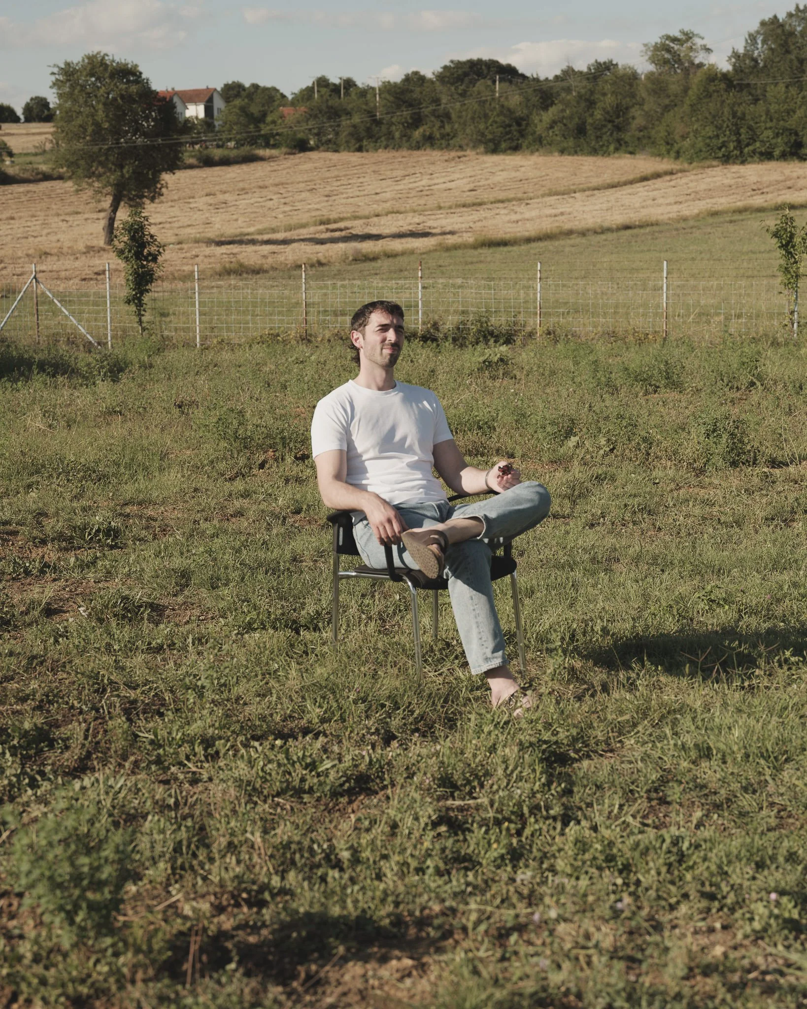 Ein junger Mann sitzt entspannt im Gras im Freien, mit einem Feld und Bäumen im Hintergrund, bei Sonnenlicht.