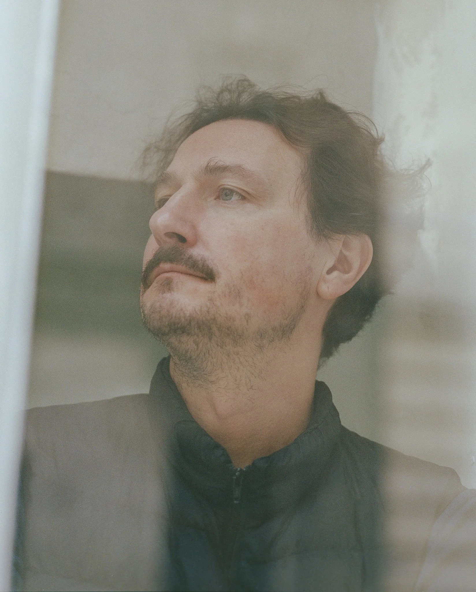 A man with dark, curly hair and a beard, wearing a black zip-up hoodie, looking to the side in front of a mirror.