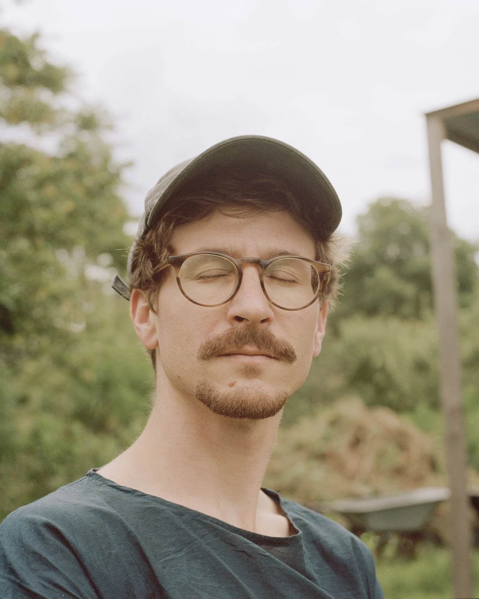 Nahaufnahme eines Mannes mit Brille, Bart und Schnurrbart, trägt eine Kapuze, steht im Freien mit Bäumen im Hintergrund.