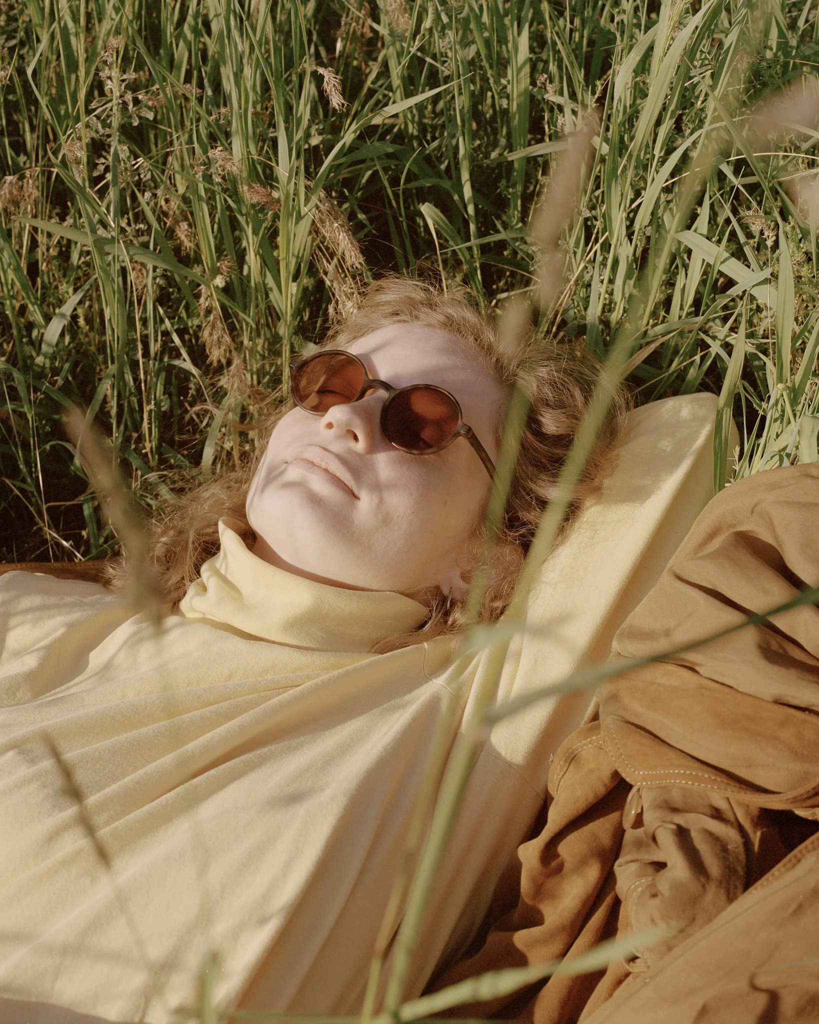 A person with light skin, wearing sunglasses and a light-colored shirt, lying among tall green grass or plants.