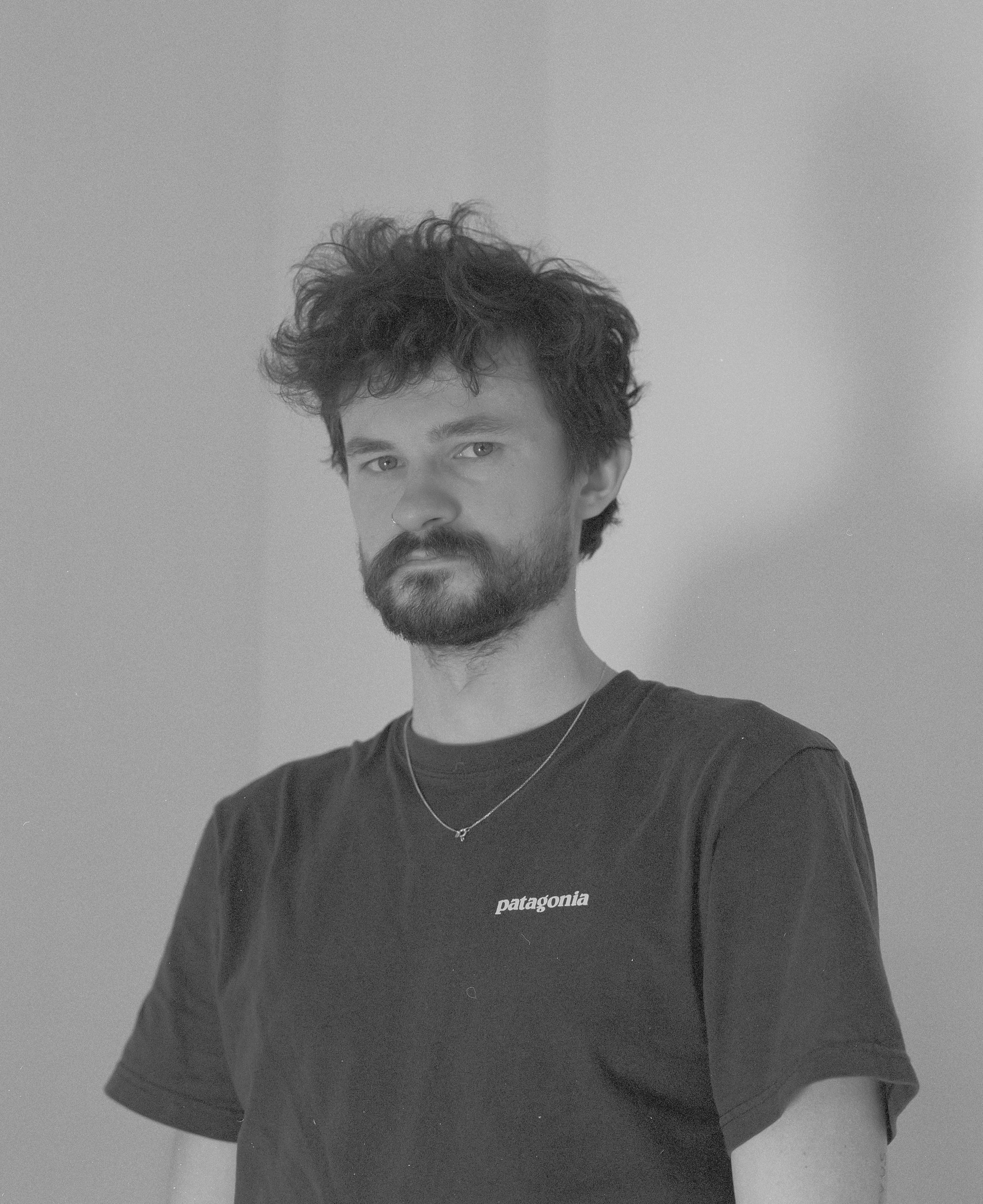 Schwarz-weiß-Fotografie eines Mannes mit lockigem Haar, Vollbart und einem schwarzen Patagonia-T-Shirt, der sich in die Kamera lehnt.