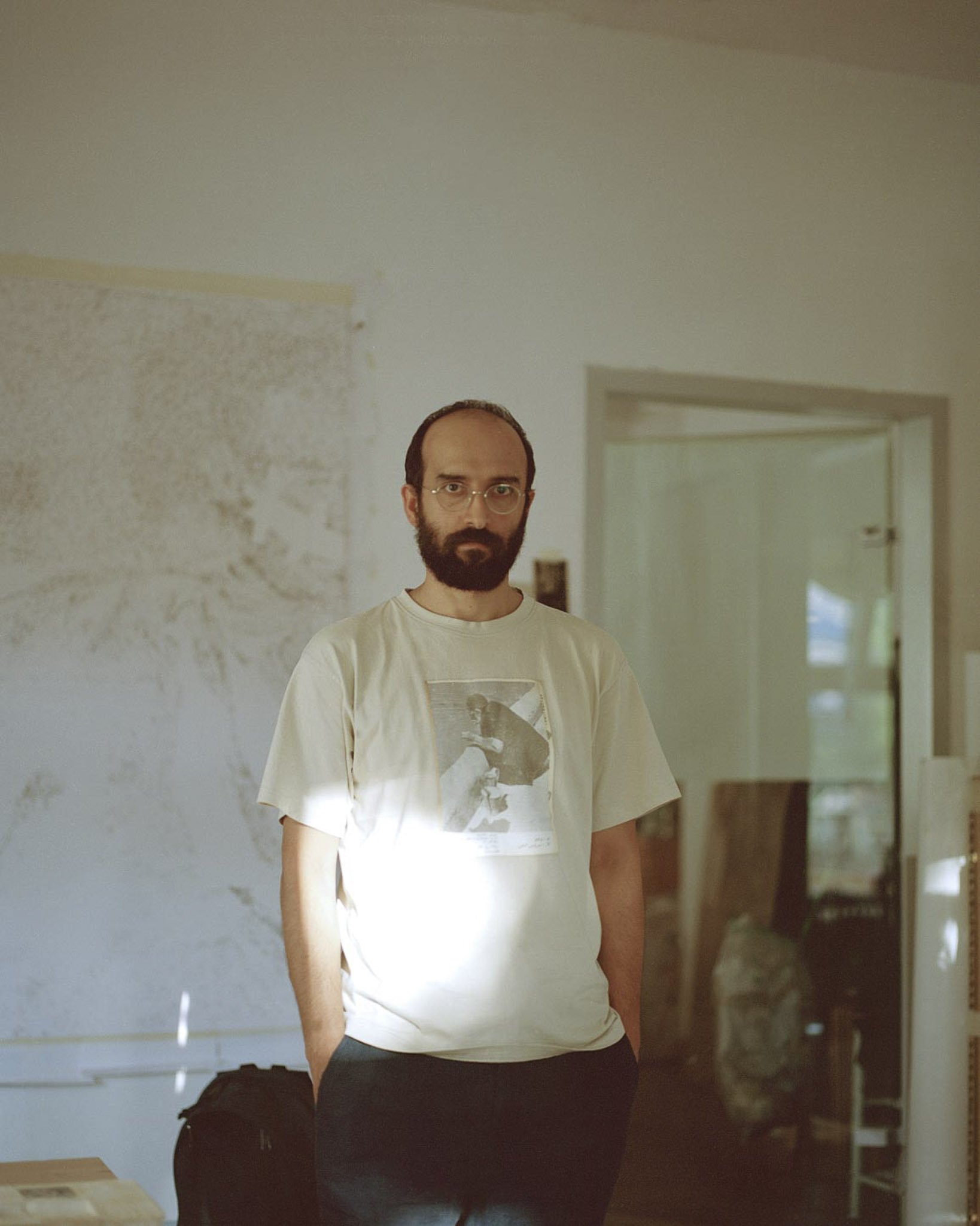 Ein Mann mit Brille, dunklem Bart und kurzen Haaren steht in einem Raum vor einer weißen Wand. Die Tür im Hintergrund ist halb offen, draußen ist bei Tageslicht. Der Mann trägt ein weißes T-Shirt mit einem Schwarz-Weiß-Bild auf der Vorderseite.