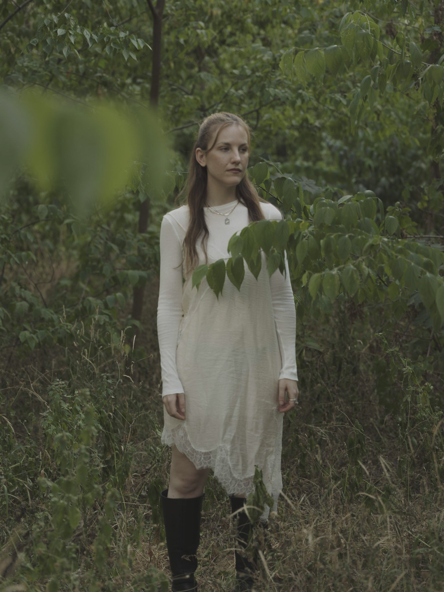 Junge Frau in weißem Kleid und schwarzen Stiefeln steht in einem grünen Wald, umgeben von Blättern und Zweigen.