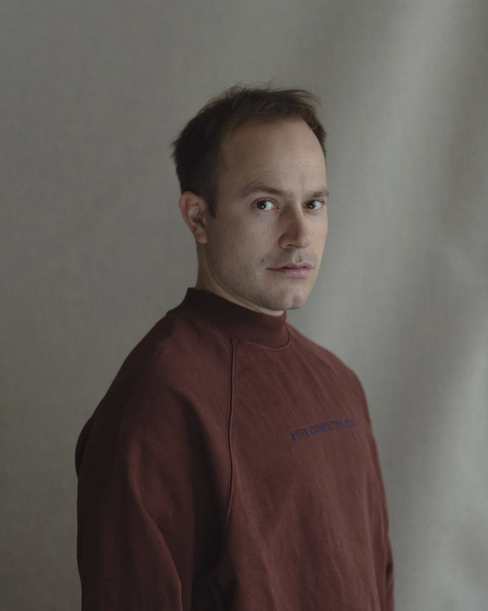 Ein junger Mann mit braunen Haaren trägt einen roten Pullover und blickt in die Kamera. Der Hintergrund ist grau.